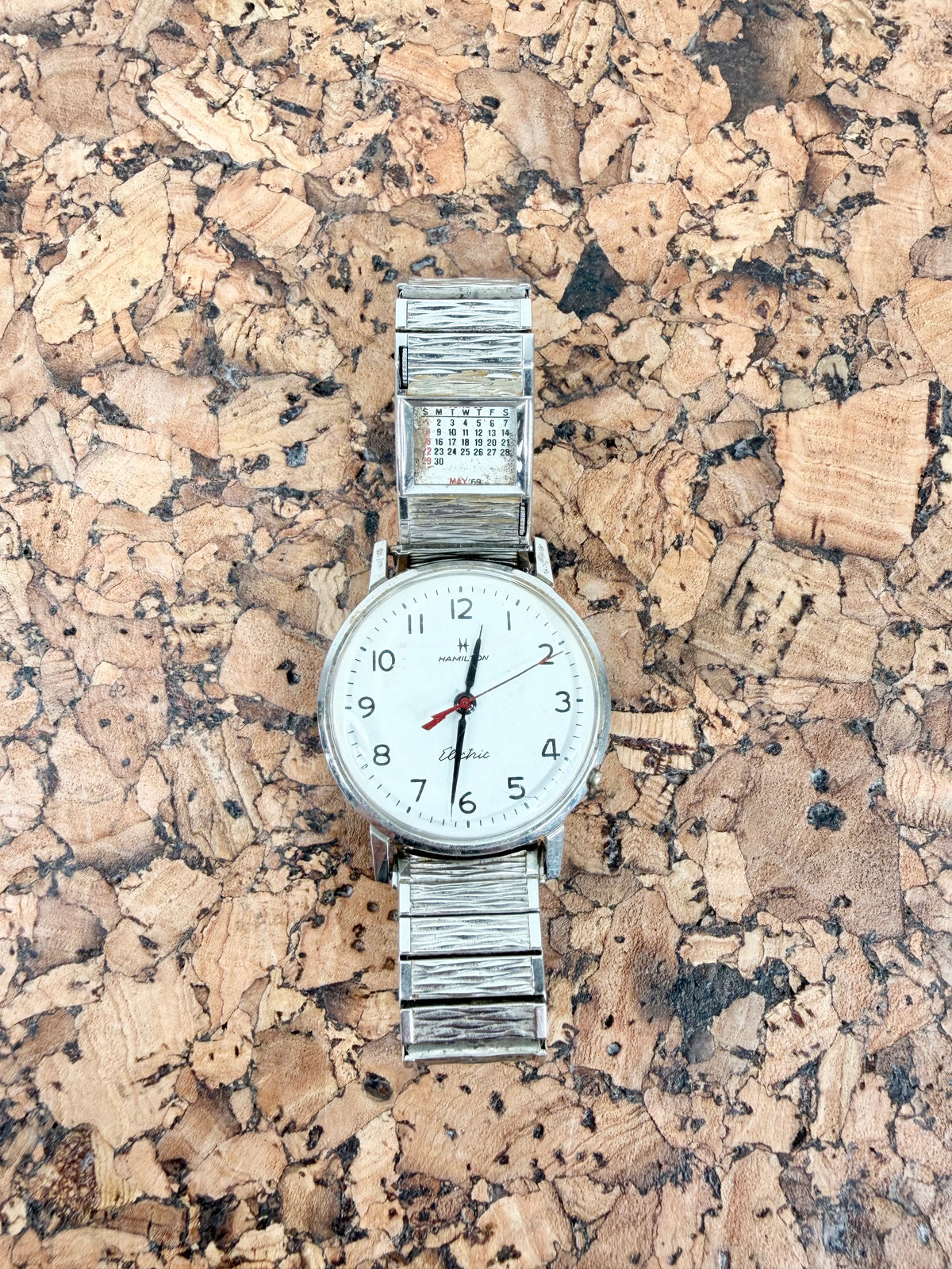 A vintage-style wristwatch with a silver-toned metal link bracelet and a white dial, placed on a textured cork surface.