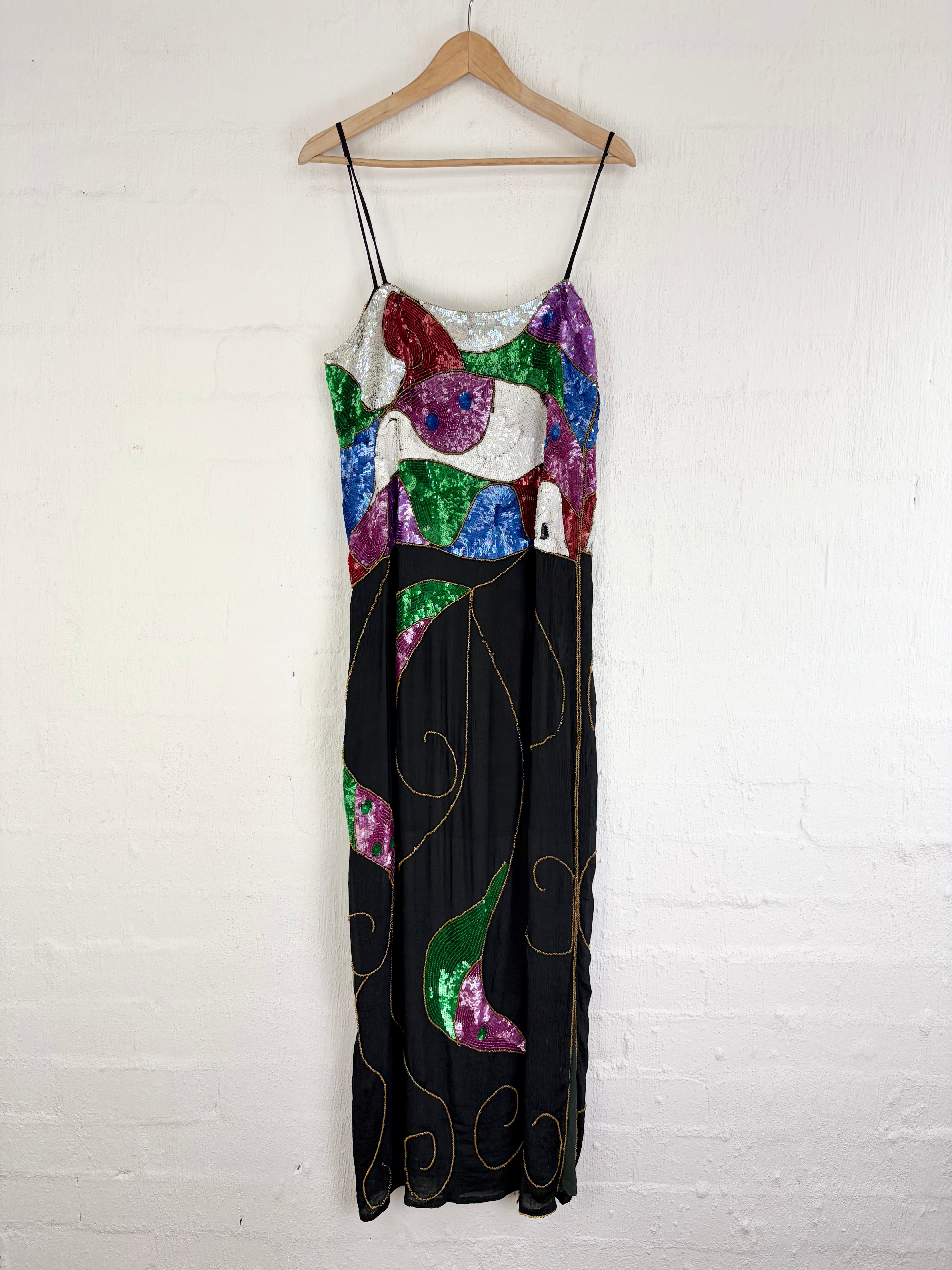 A sleeveless maxi dress with thin black spaghetti straps, featuring a colorful abstract patchwork print on a black base, hanging on a wooden hanger against a white painted brick wall.