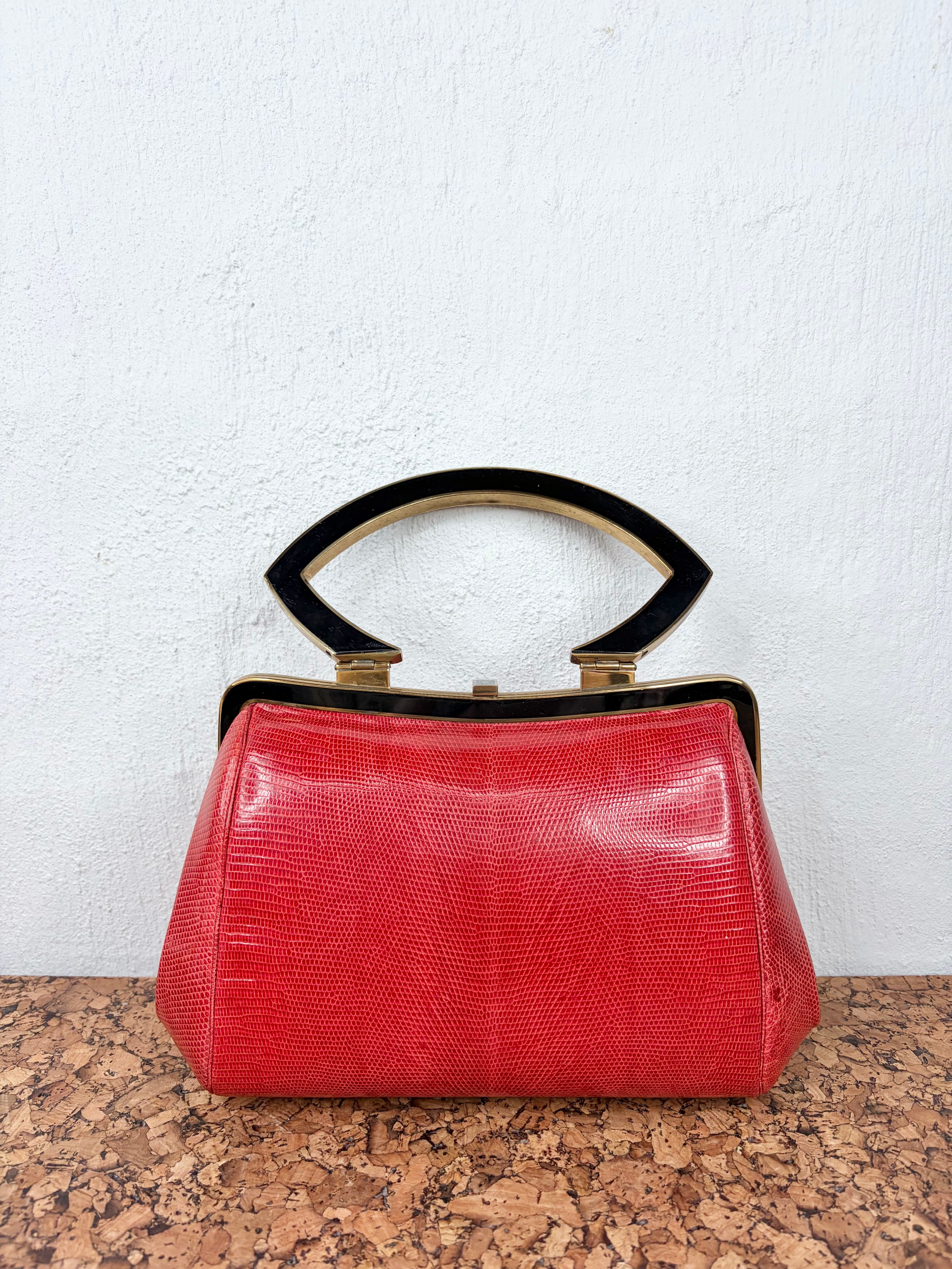 A vintage red python-textured handbag with a black arched handle and gold-tone hardware, resting on a cork surface against a white wall.