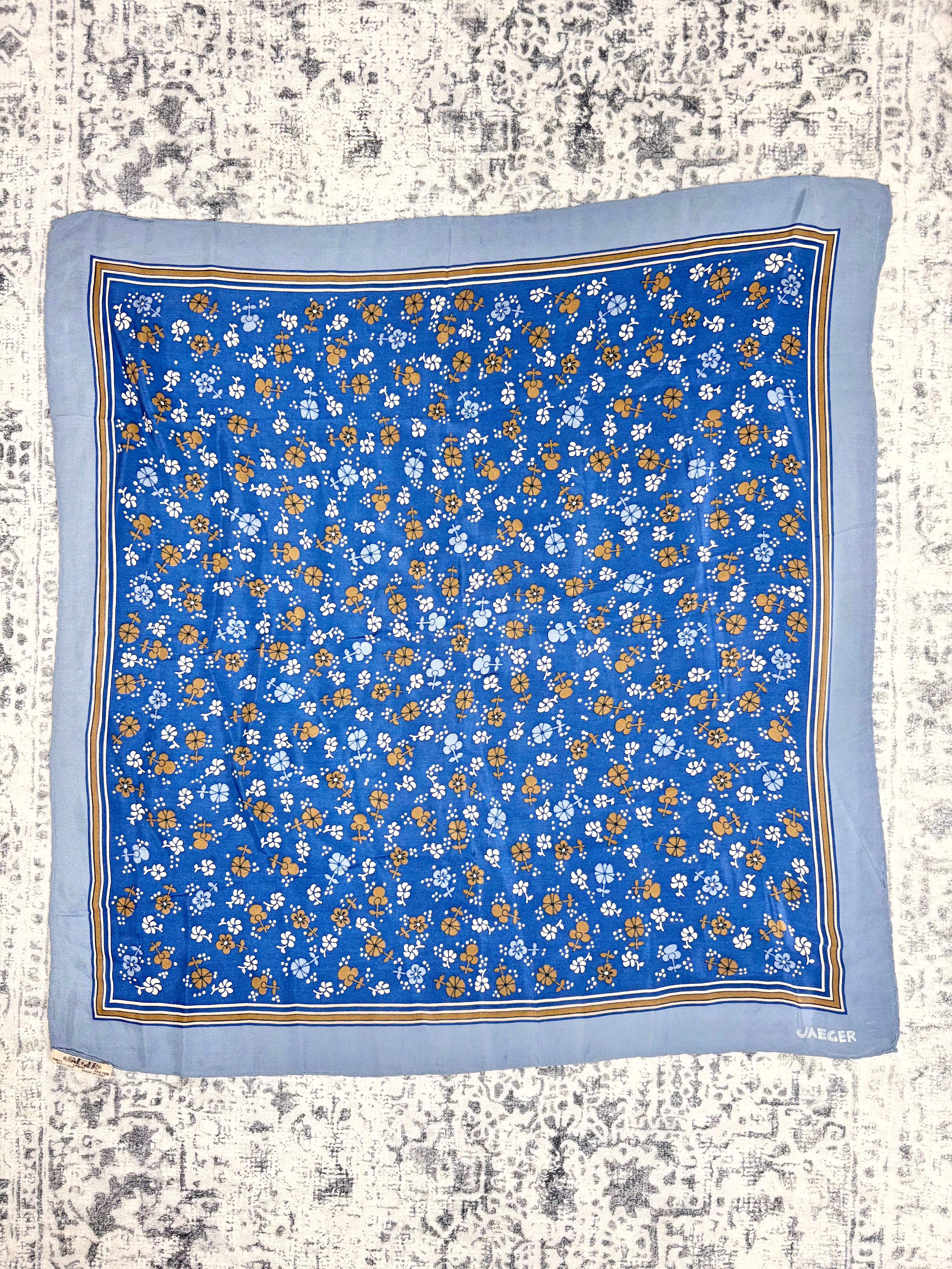 A square blue scarf with a dense small floral print in white and orange, finished with a light blue border and thin gold stripes, laid flat on a distressed off-white background. The brand name 'JACOBS' is visible in the bottom right corner.