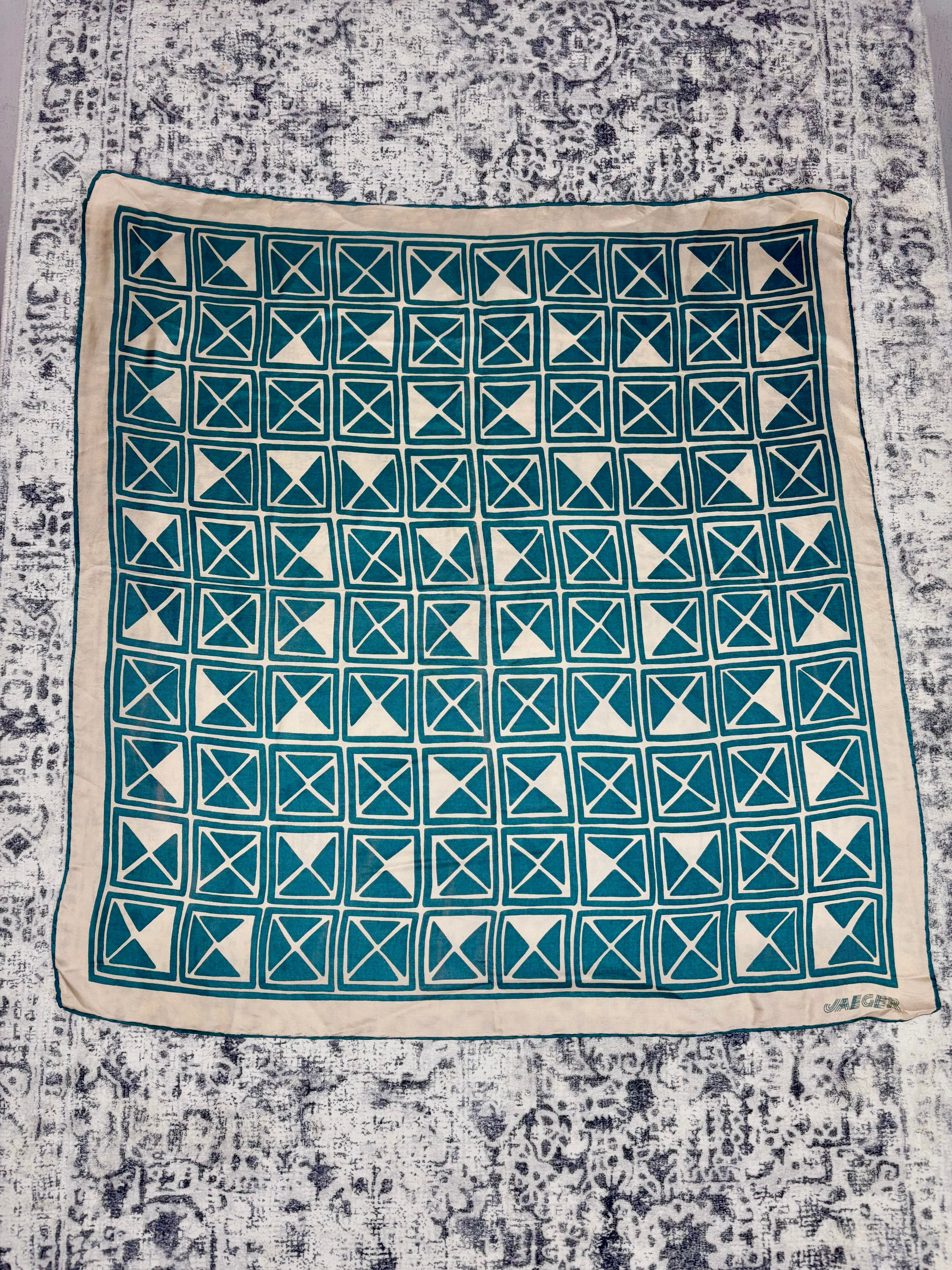 A square textile with a teal and cream geometric block-print pattern, laid flat on a distressed gray and white patterned rug.