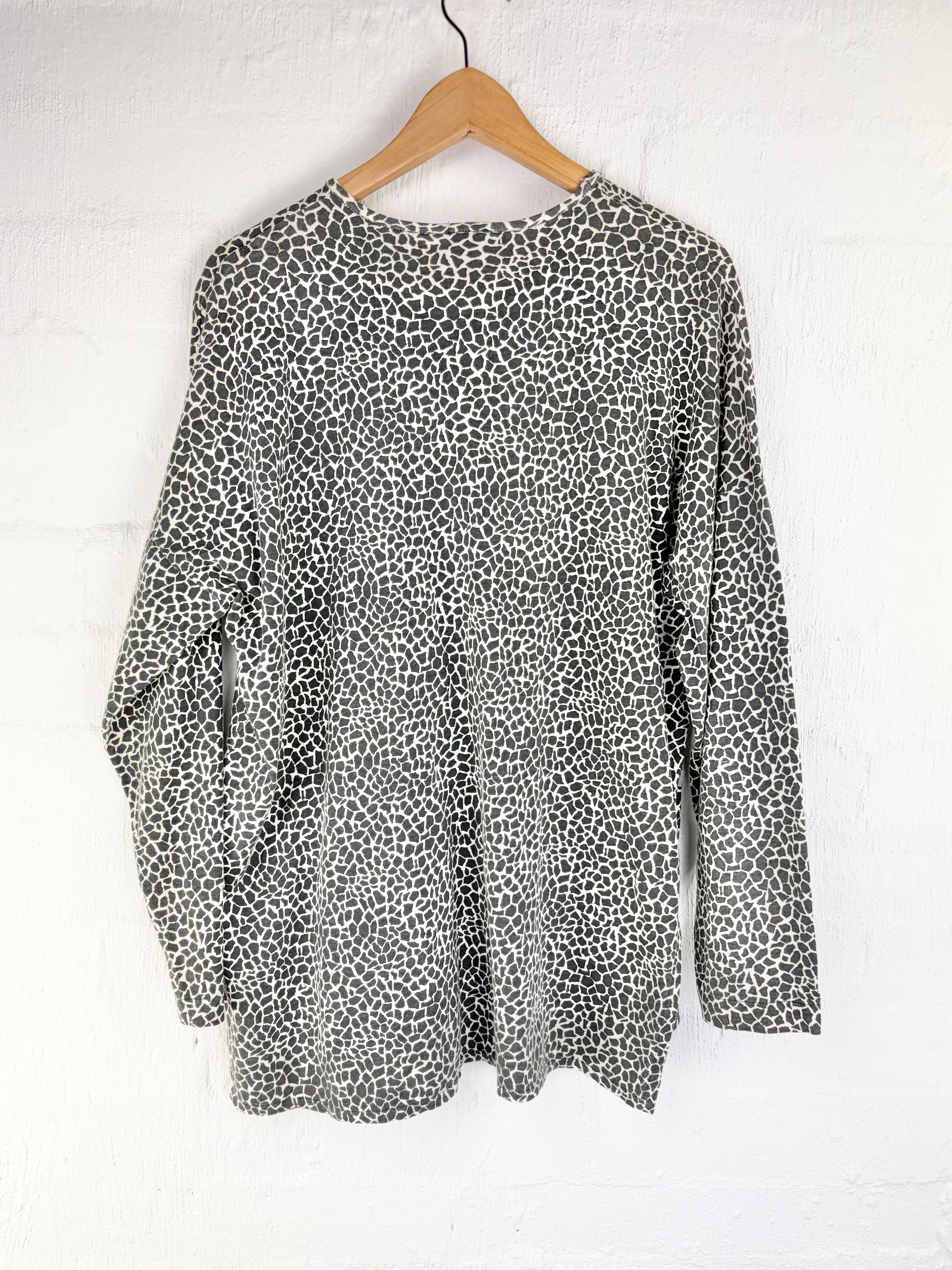 A long-sleeve top with an all-over black and white leopard print pattern, hanging on a wooden hanger against a white painted brick wall.