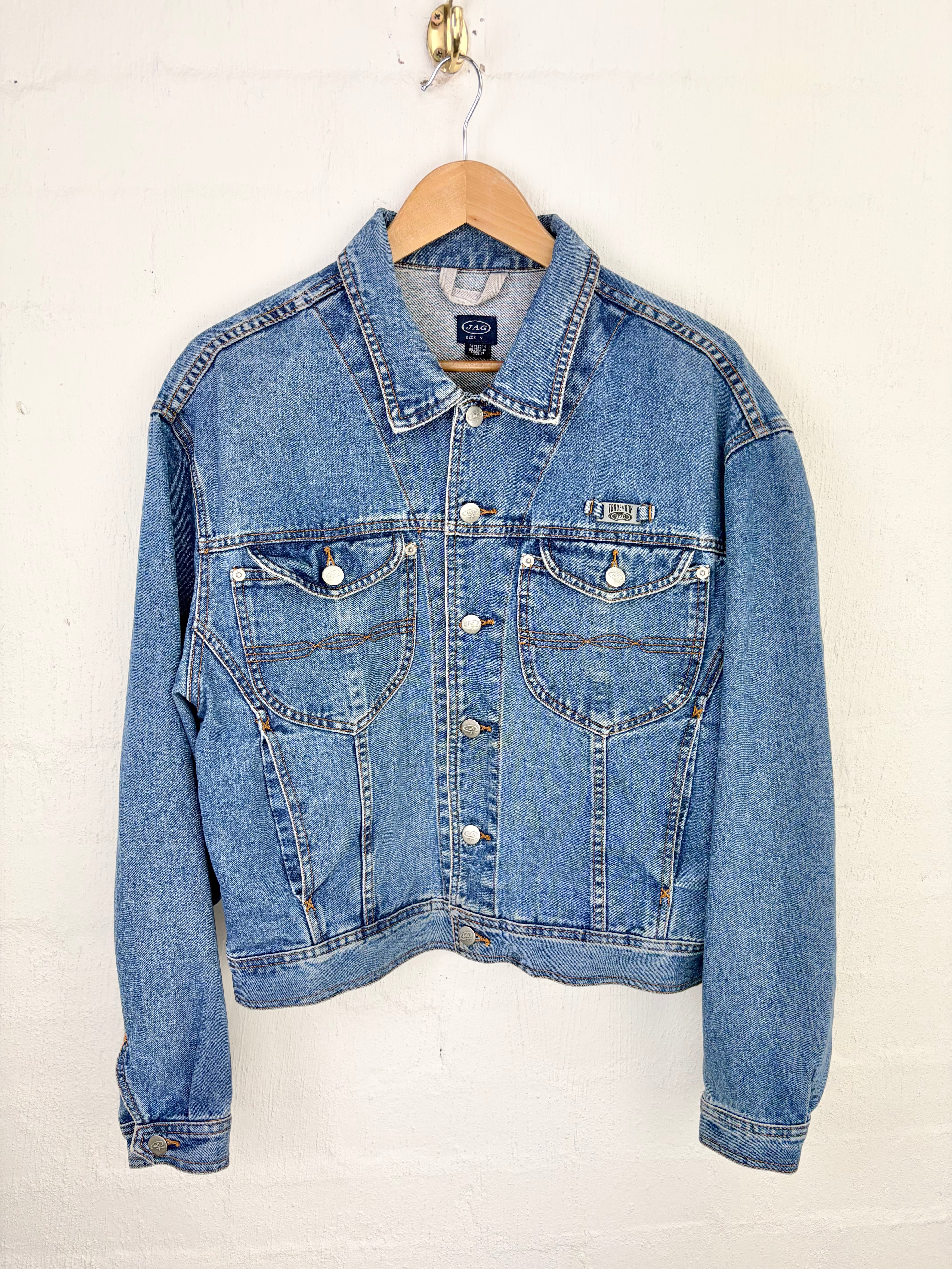 A light blue denim jacket hanging on a wooden hanger against a white painted brick wall. The jacket features a classic collar, button-up front, and two buttoned chest pockets.