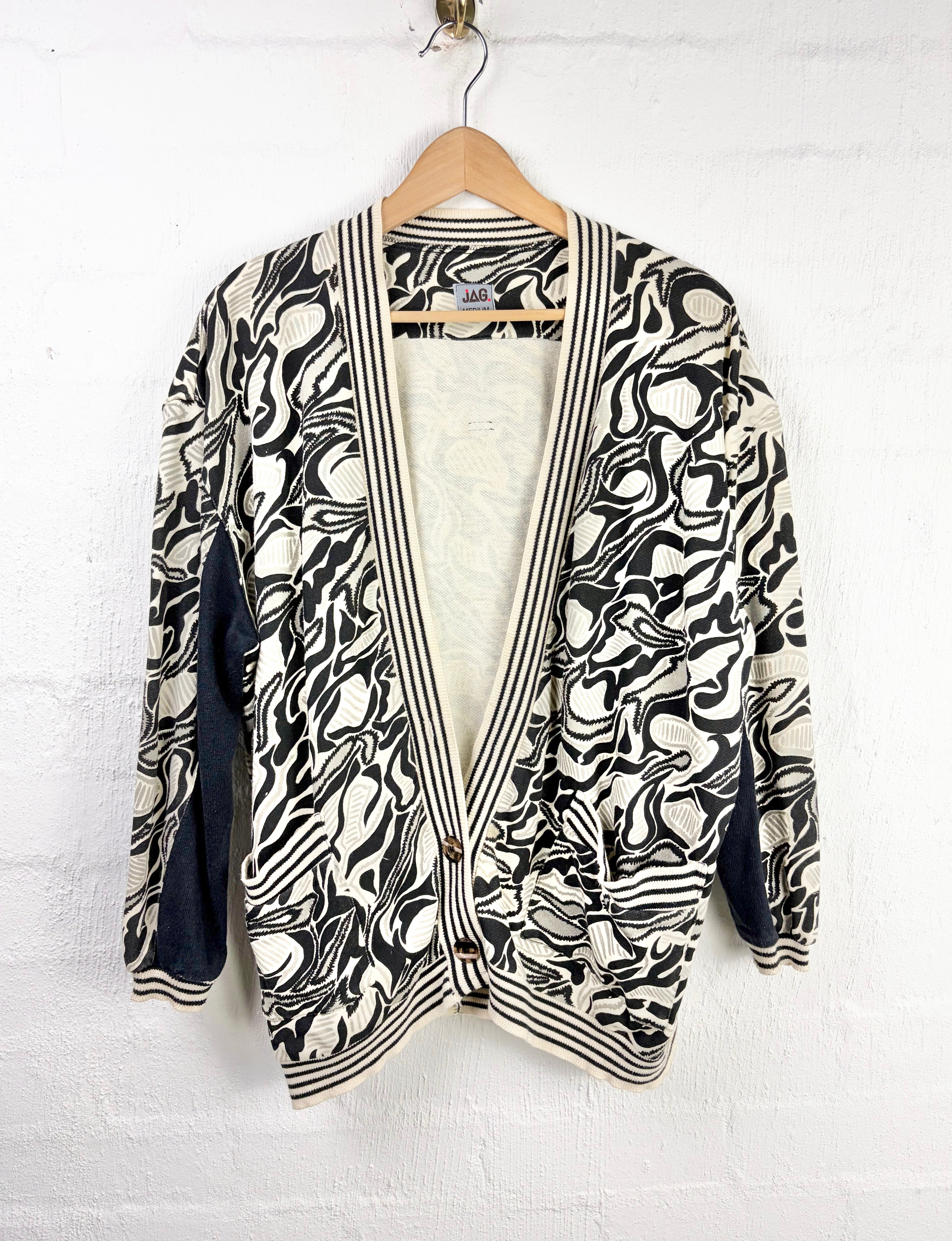 A black and off-white abstract patterned cardigan with dark navy blue contrast panels on the sleeves and striped trim, hanging on a wooden hanger against a white brick wall.