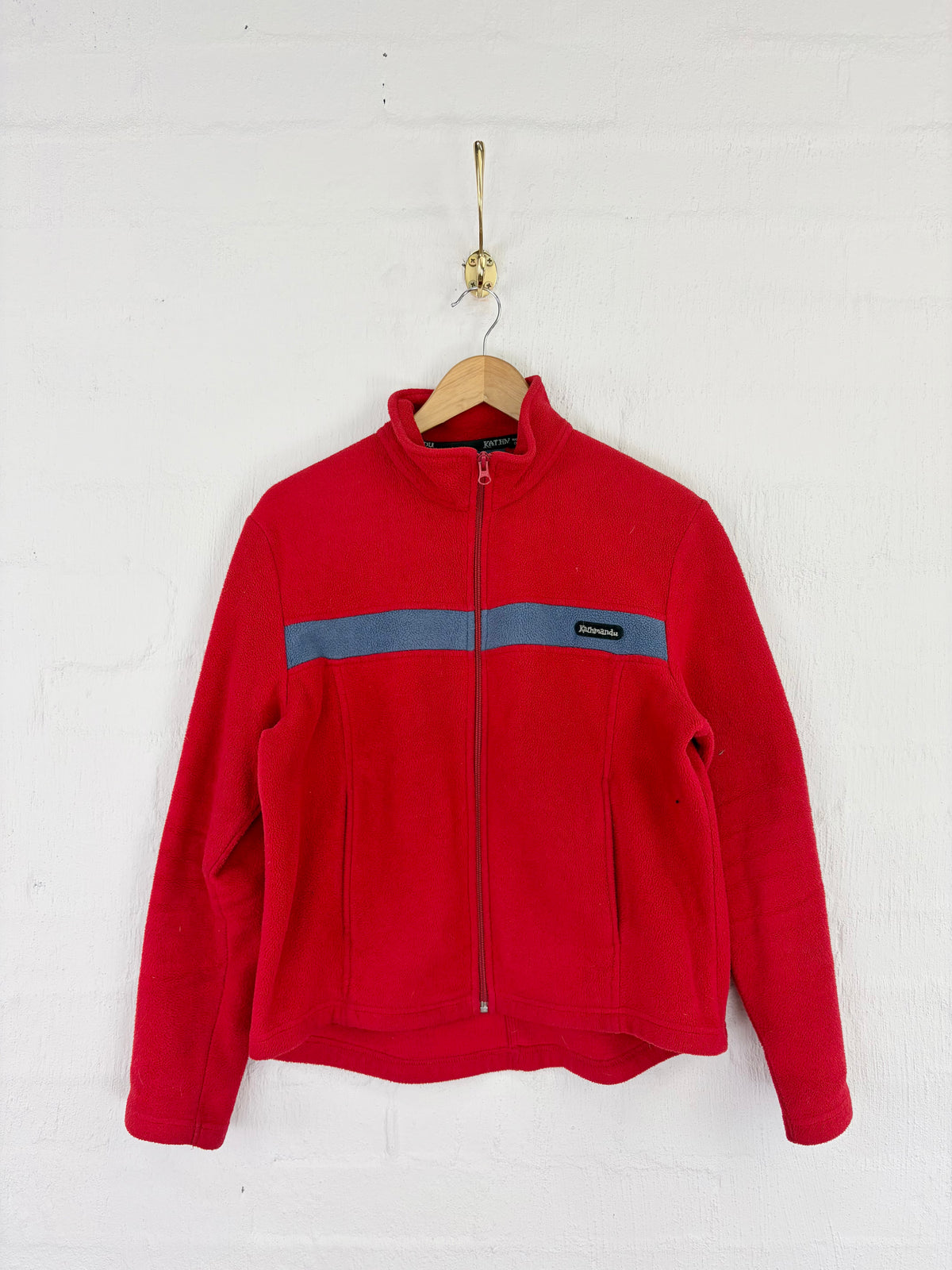 A red fleece zip-up jacket with a gray horizontal chest stripe, hanging on a wooden hanger against a white painted brick wall.