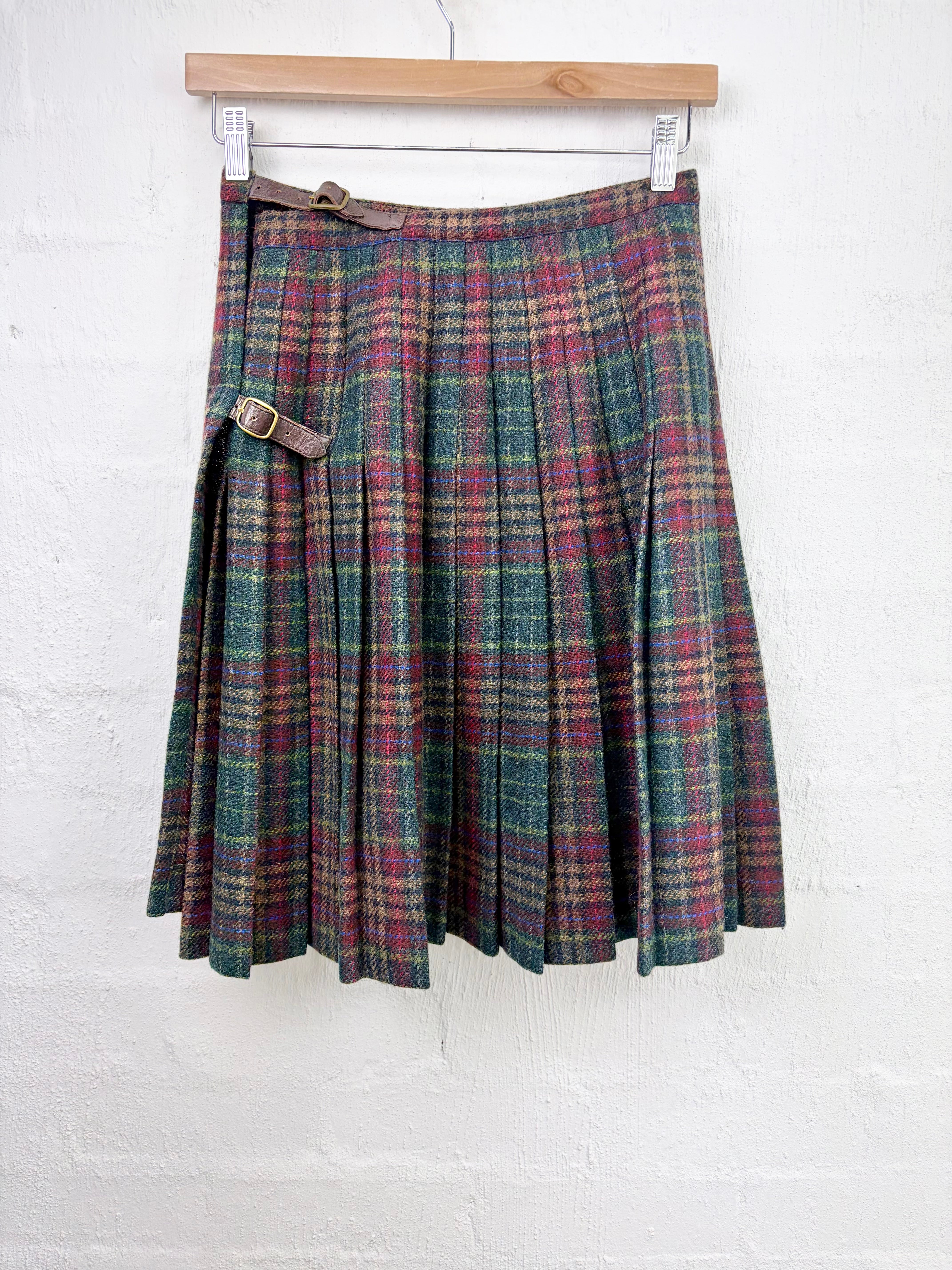 A pleated plaid skirt in green, red, and beige tartan, hanging from a wooden hanger against a white painted brick wall. It features a brown leather belt with a gold buckle at the waist.