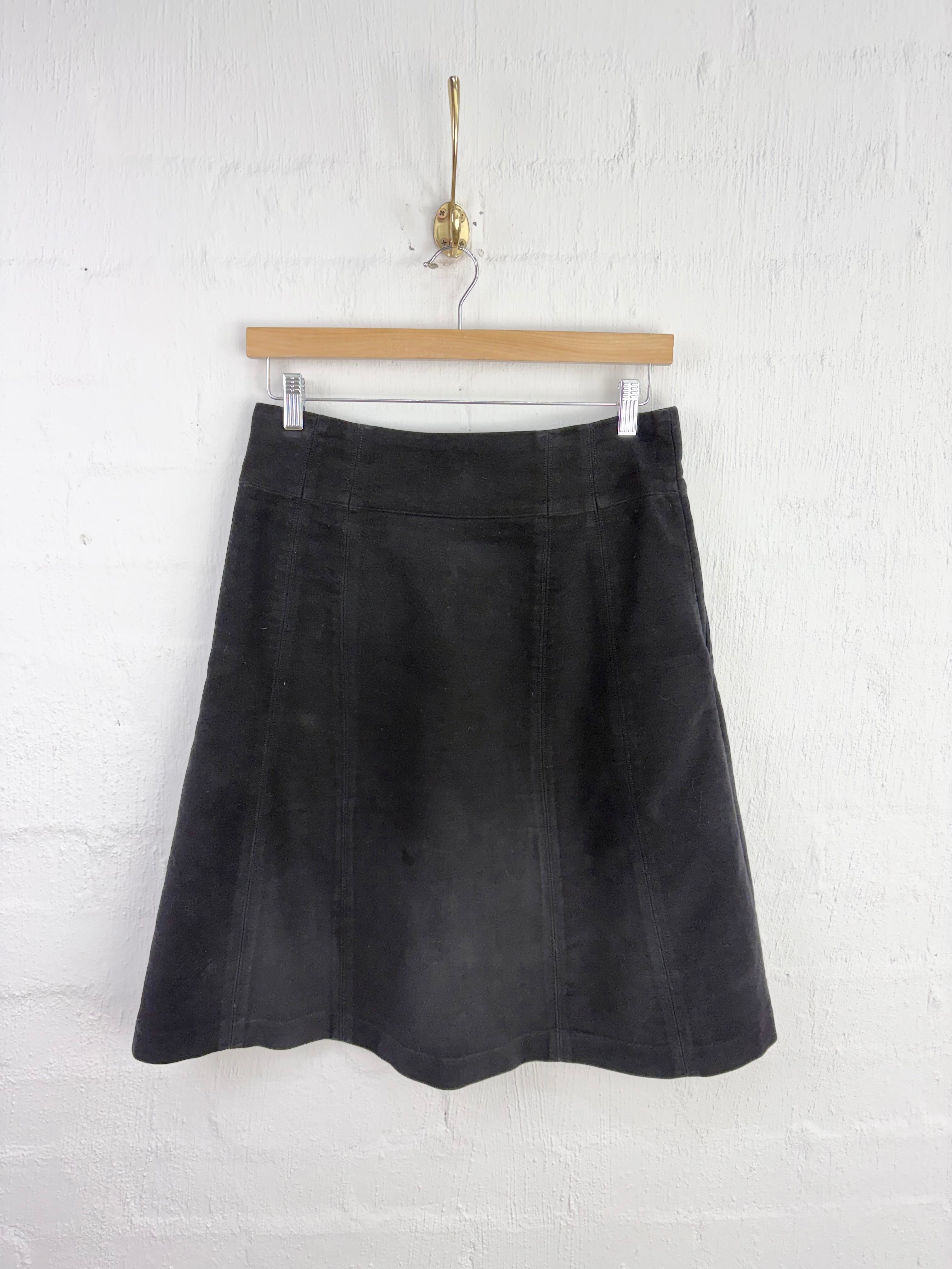 A black A-line skirt made of a textured fabric, hanging from a wooden hanger against a white painted brick wall.