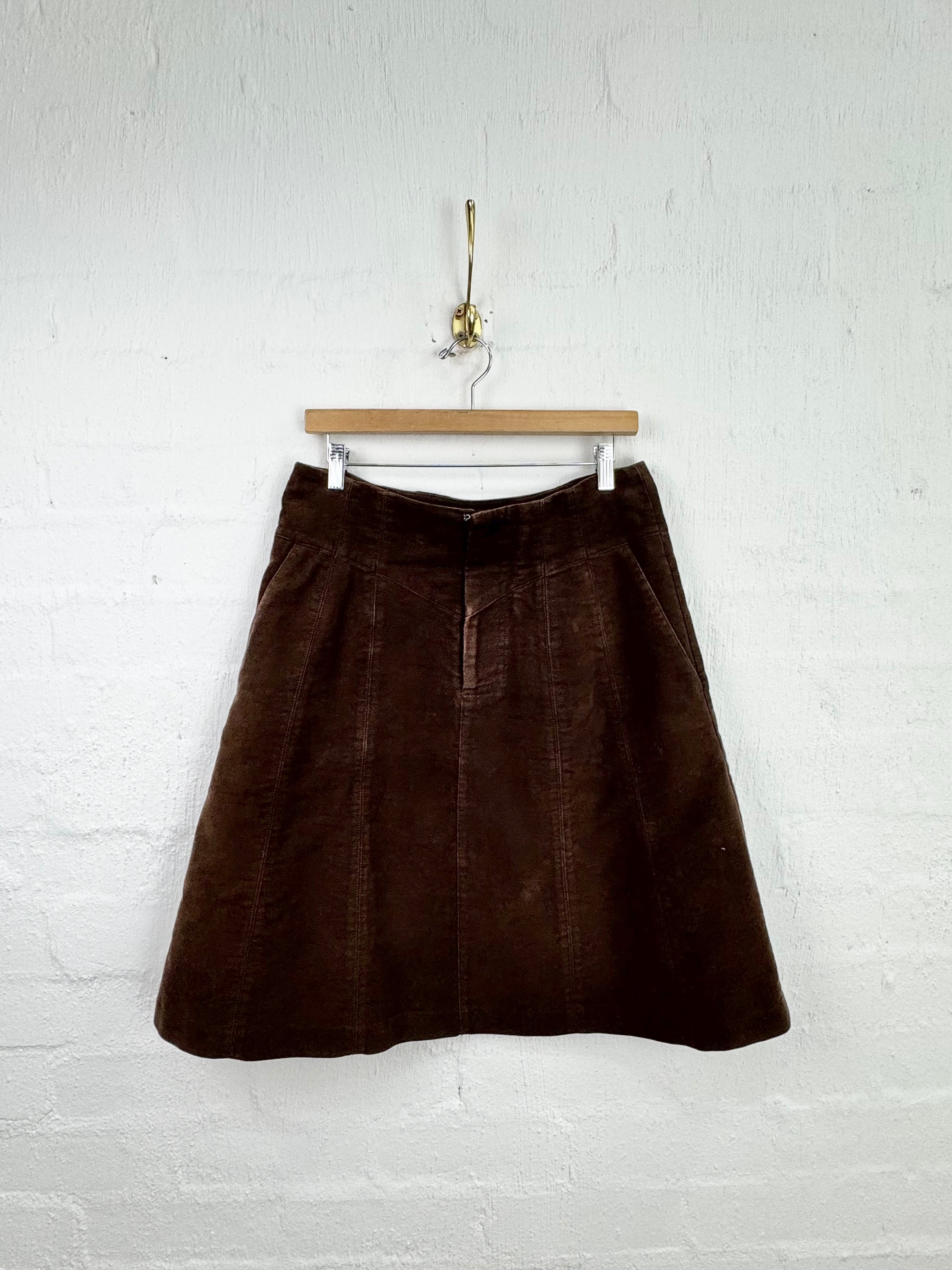 A dark brown corduroy A-line skirt hanging from a wooden hanger against a white painted brick wall.