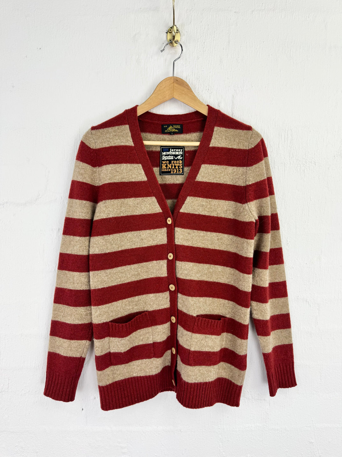 A vintage knit cardigan sweater with horizontal red and beige stripes, hanging on a wooden hanger against a white painted brick wall. The cardigan features a V-neck, button closure, and front pockets.