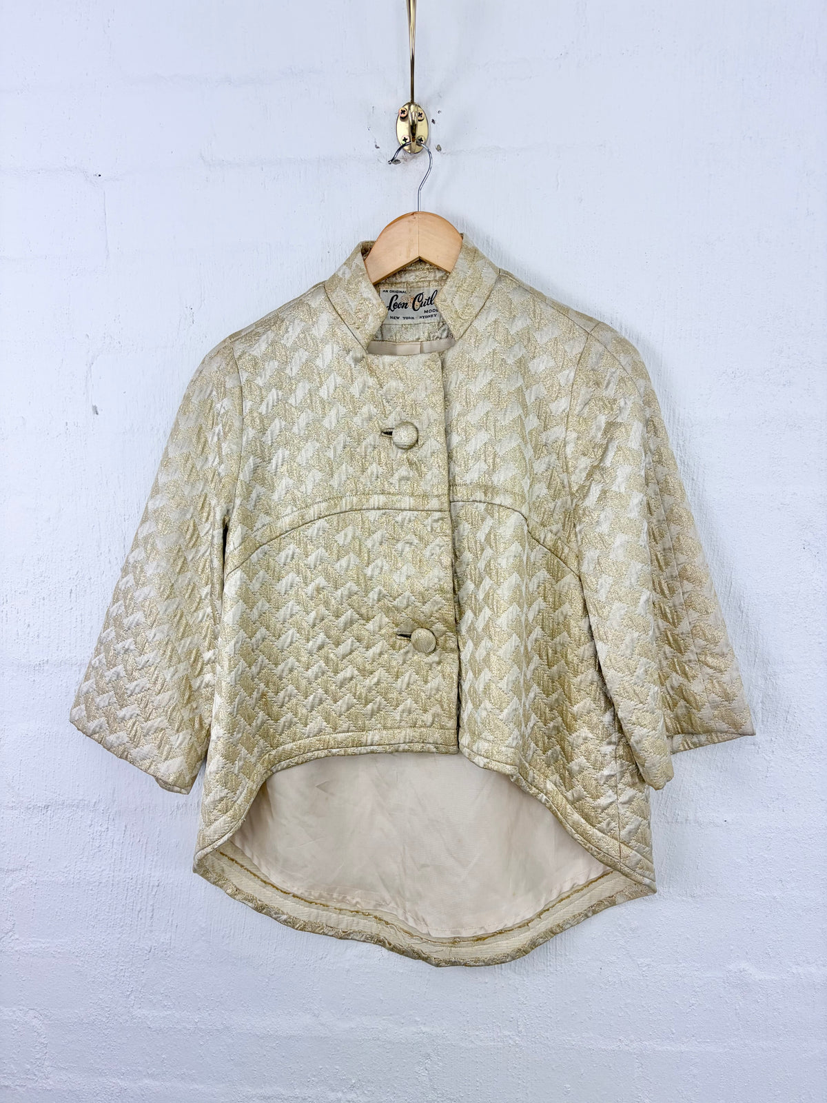 A cropped quilted jacket in a textured beige and cream jacquard pattern, hanging on a wooden hanger against a white painted brick wall.