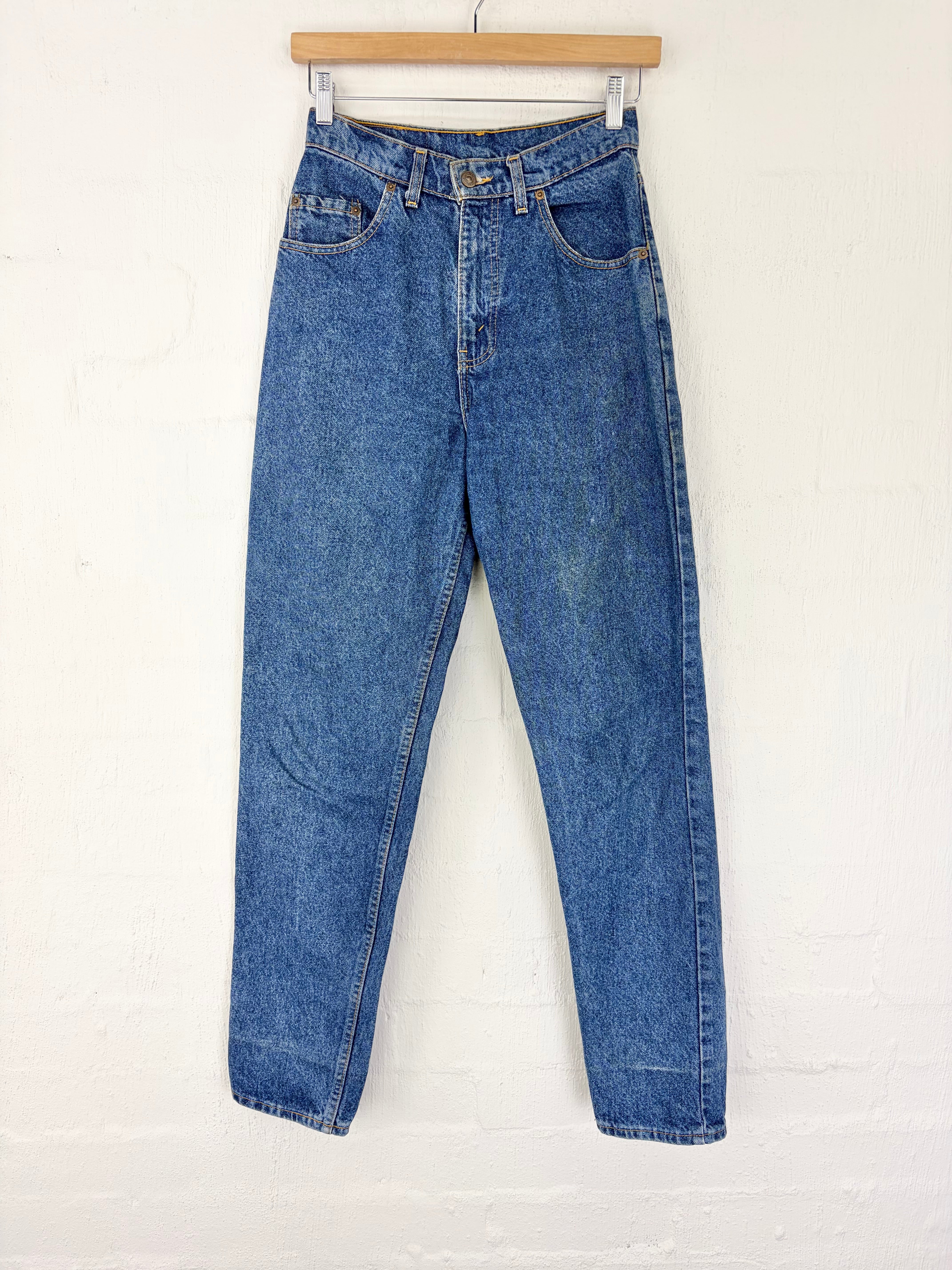 A pair of medium wash blue denim jeans hanging from a wooden hanger against a white painted brick wall.
