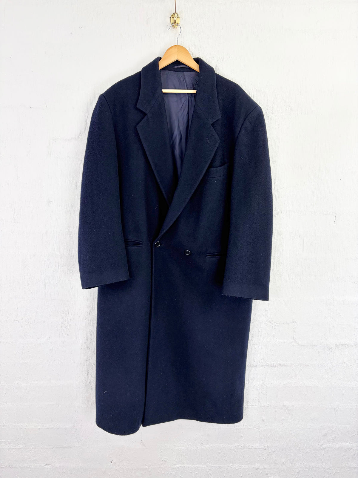 A dark navy blue wool-blend coat hanging on a wooden hanger against a white painted brick wall.