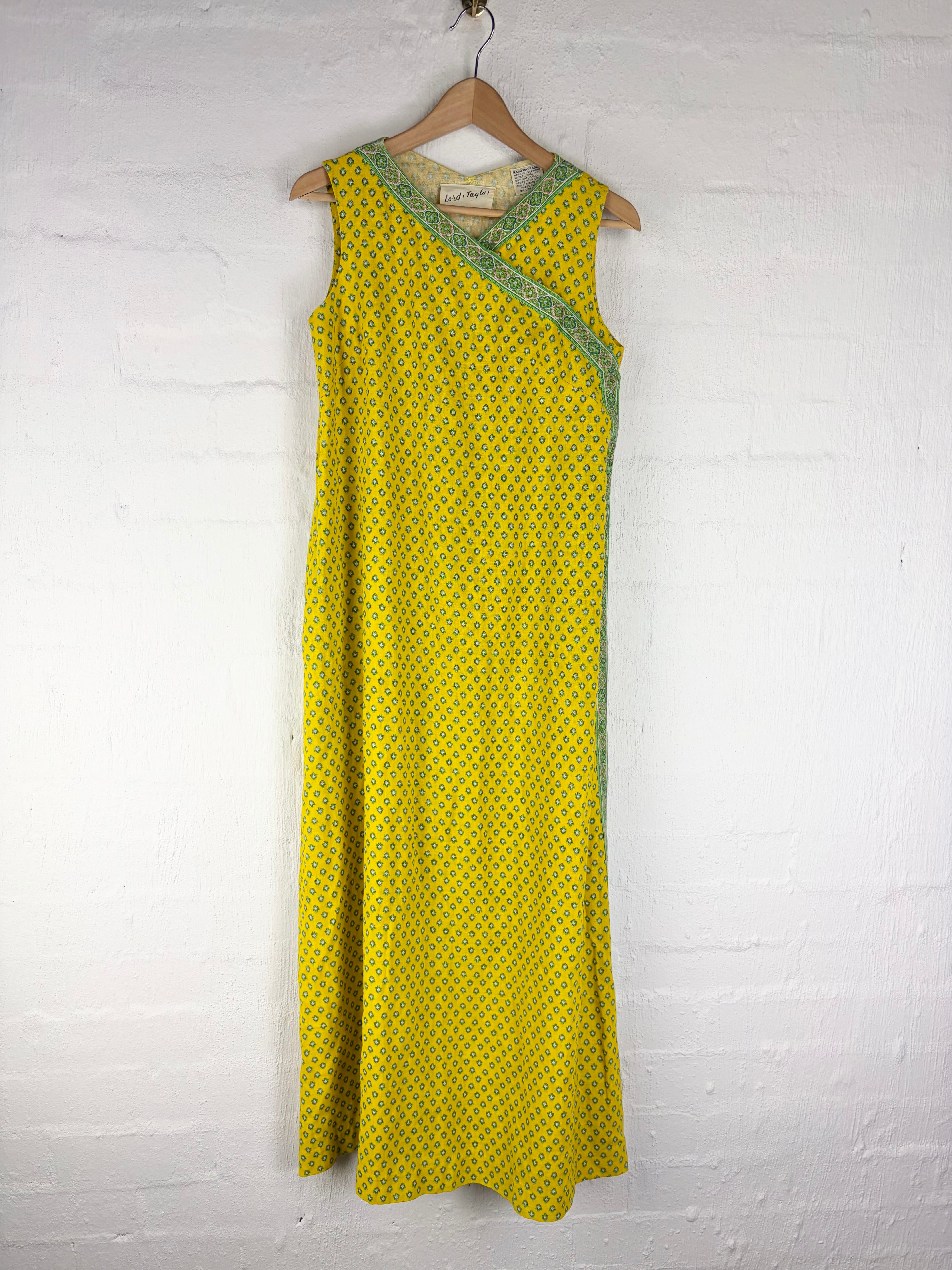 A sleeveless yellow maxi dress with a small green geometric pattern and green trim, hanging on a wooden hanger against a white brick wall.