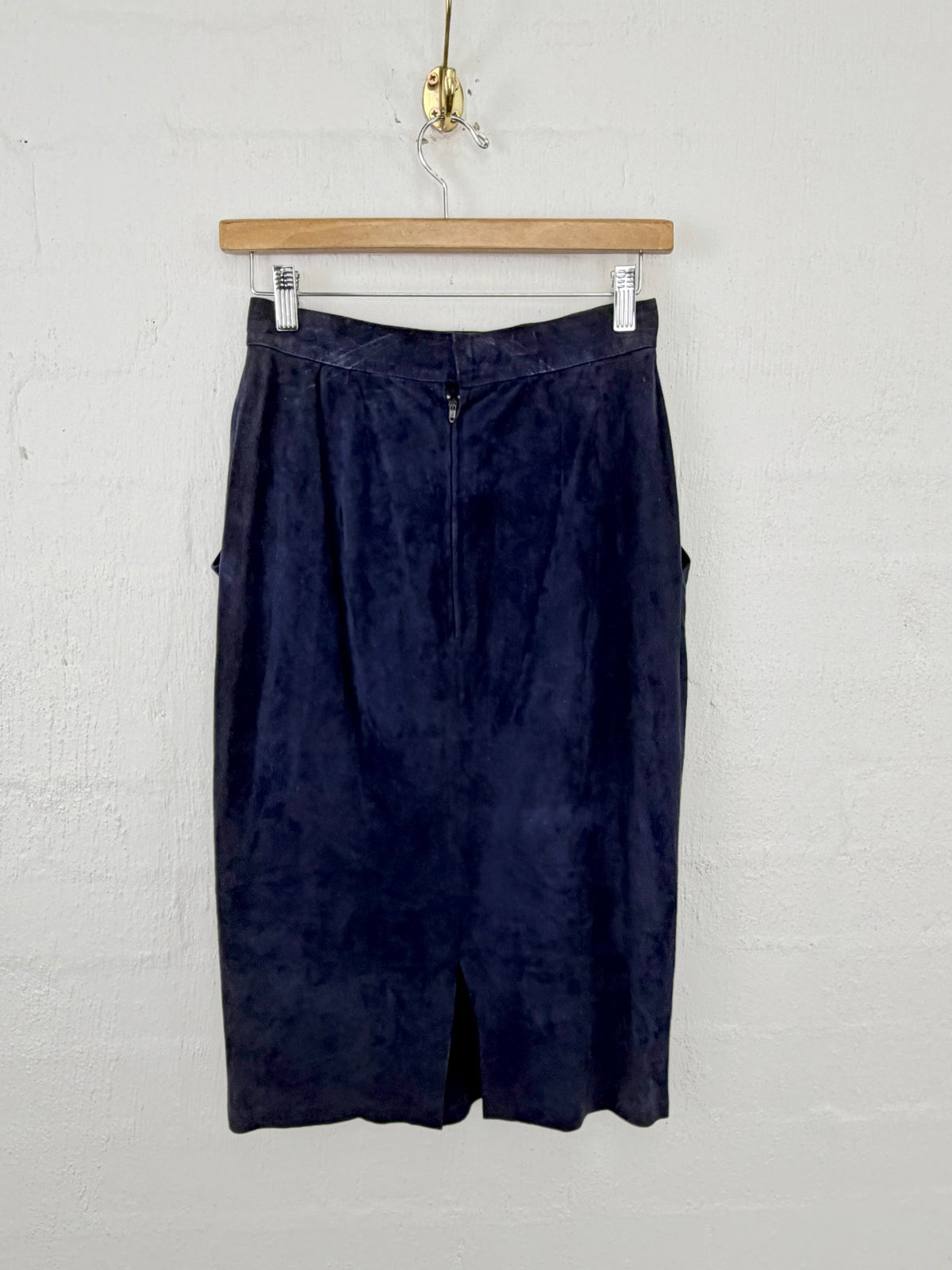 A dark navy blue velvet or velour skirt hanging from a wooden hanger against a white painted brick wall.