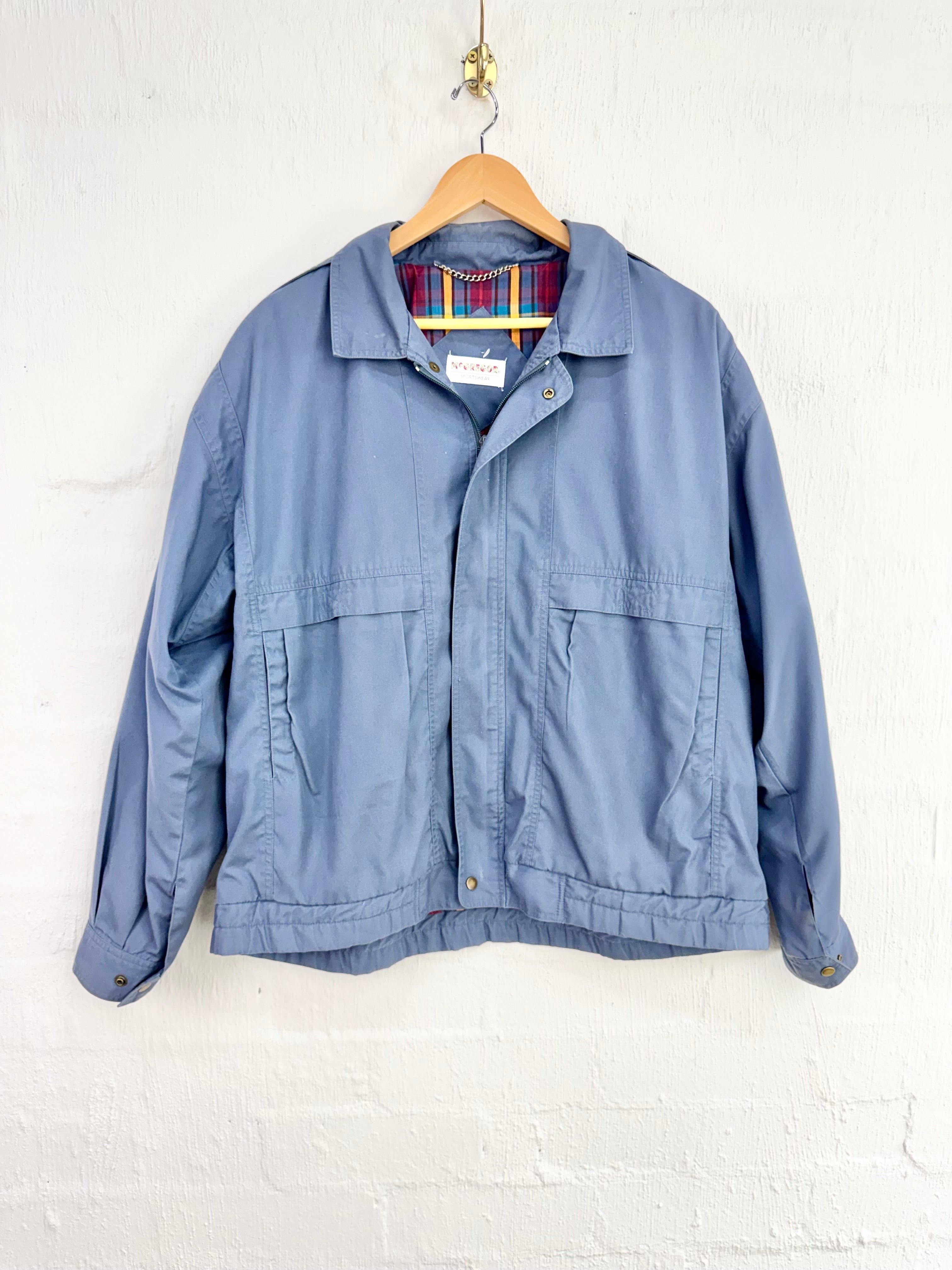 A dusty blue utility jacket with a pointed collar, button front, and two front flap pockets, hanging on a wooden hanger against a white painted brick wall.