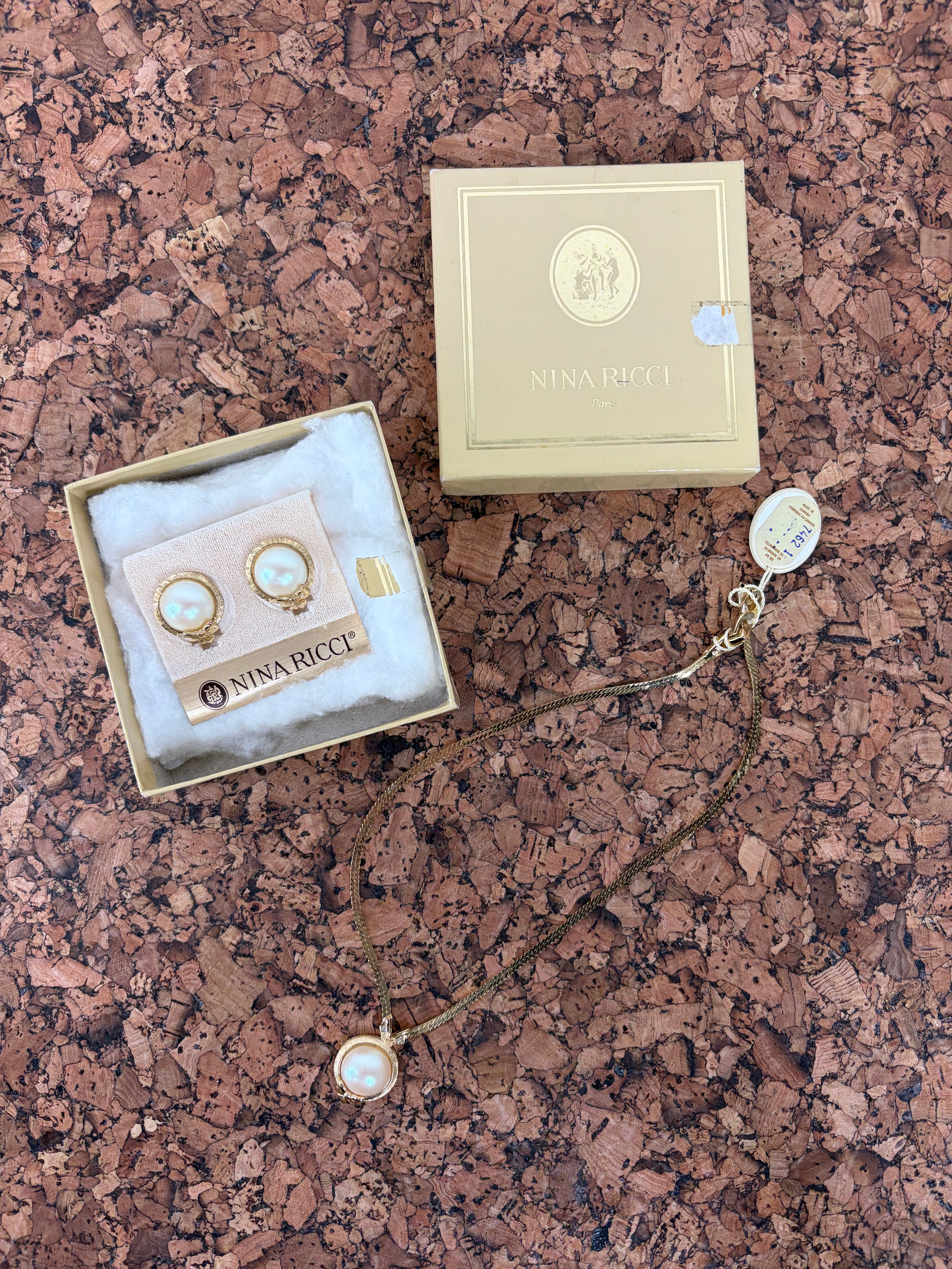 Nina Ricci jewelry set featuring a gold necklace with a white pearl pendant and matching stud earrings, displayed with their branded packaging on a textured brown cork background.
