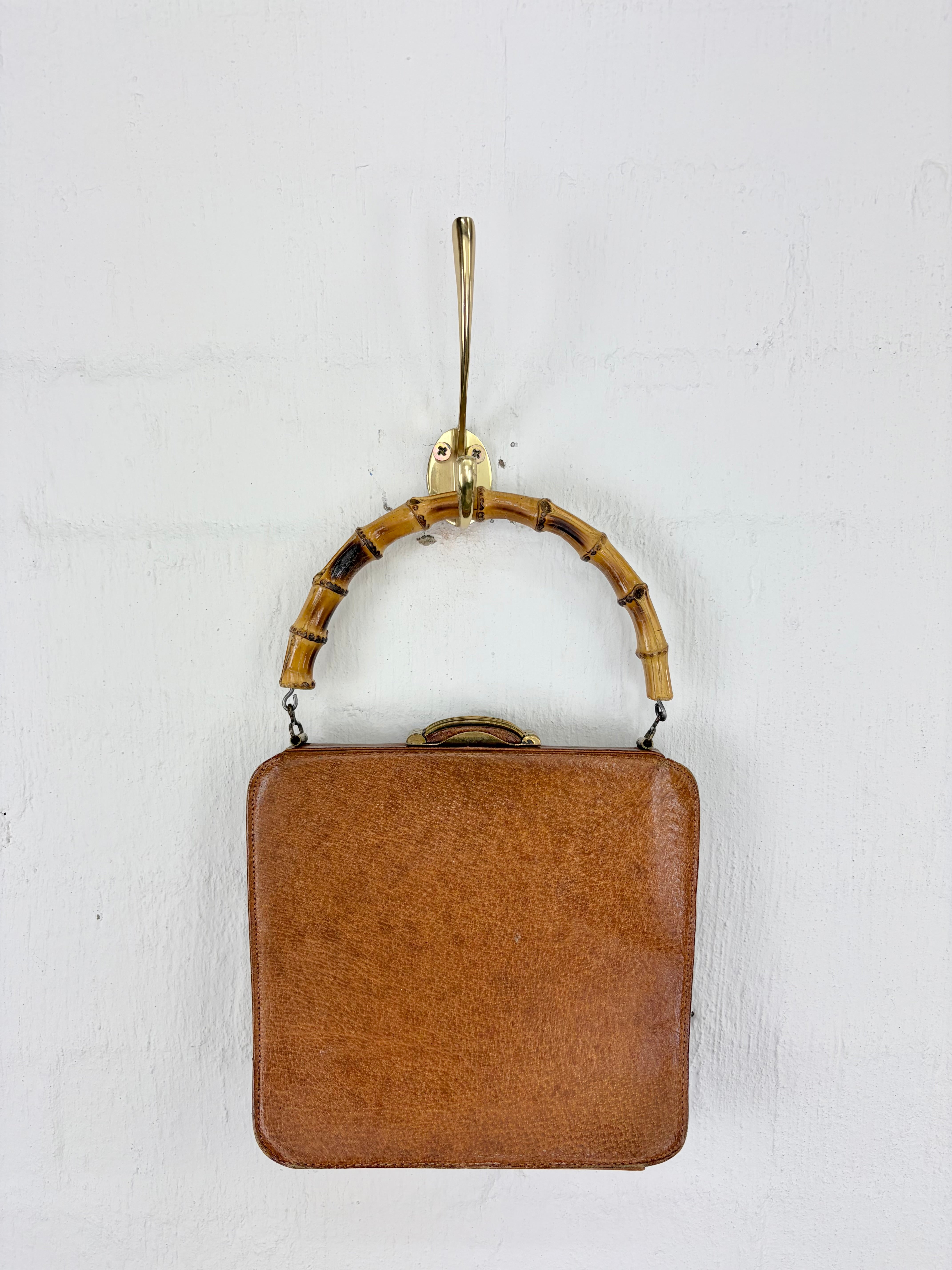 A vintage brown leather handbag with a bamboo handle and gold-tone hardware, hanging against a white textured wall.