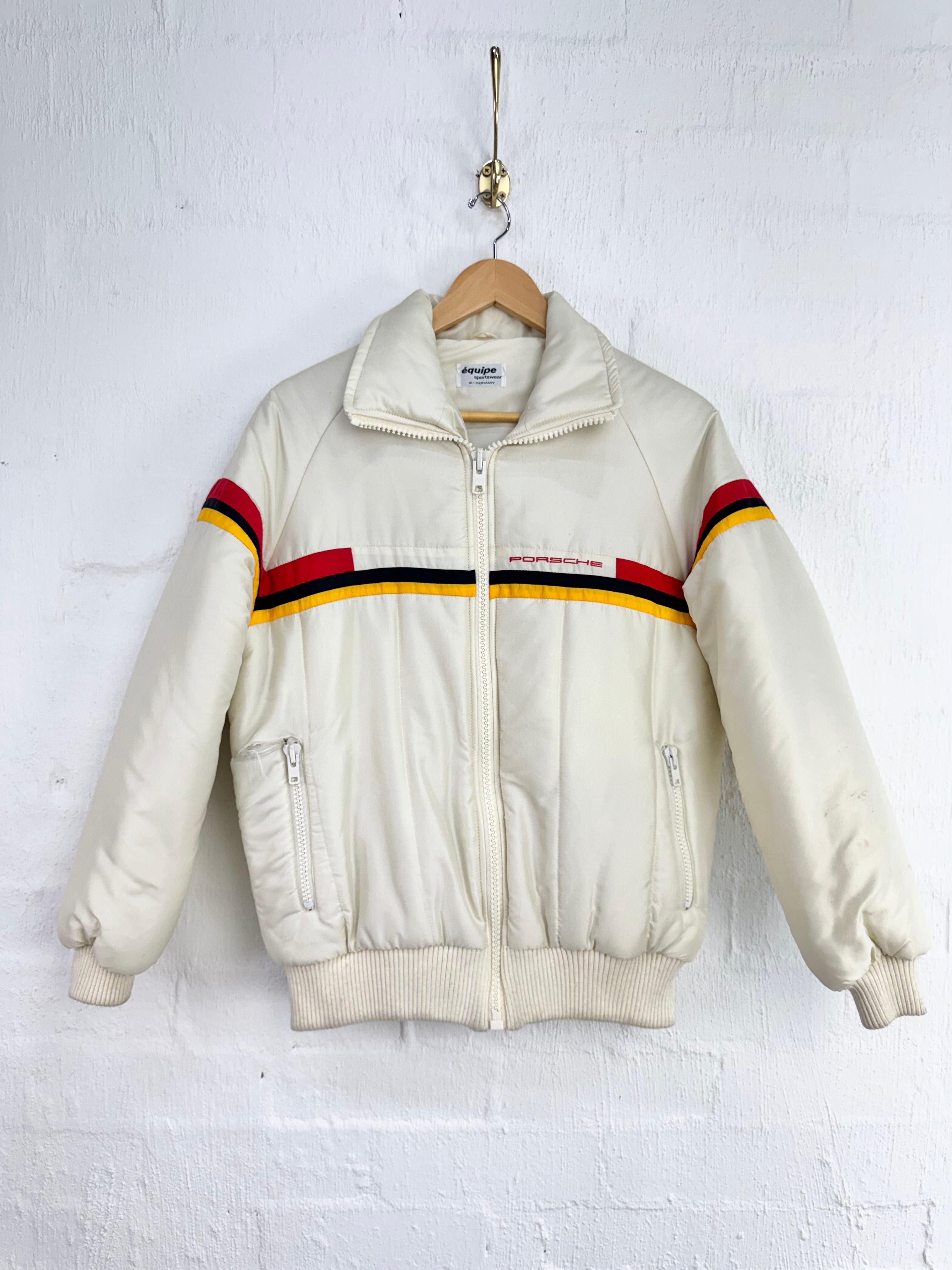 An off-white vintage bomber jacket with red, black, and yellow stripes across the chest and sleeves, hanging on a wooden hanger against a white brick wall.