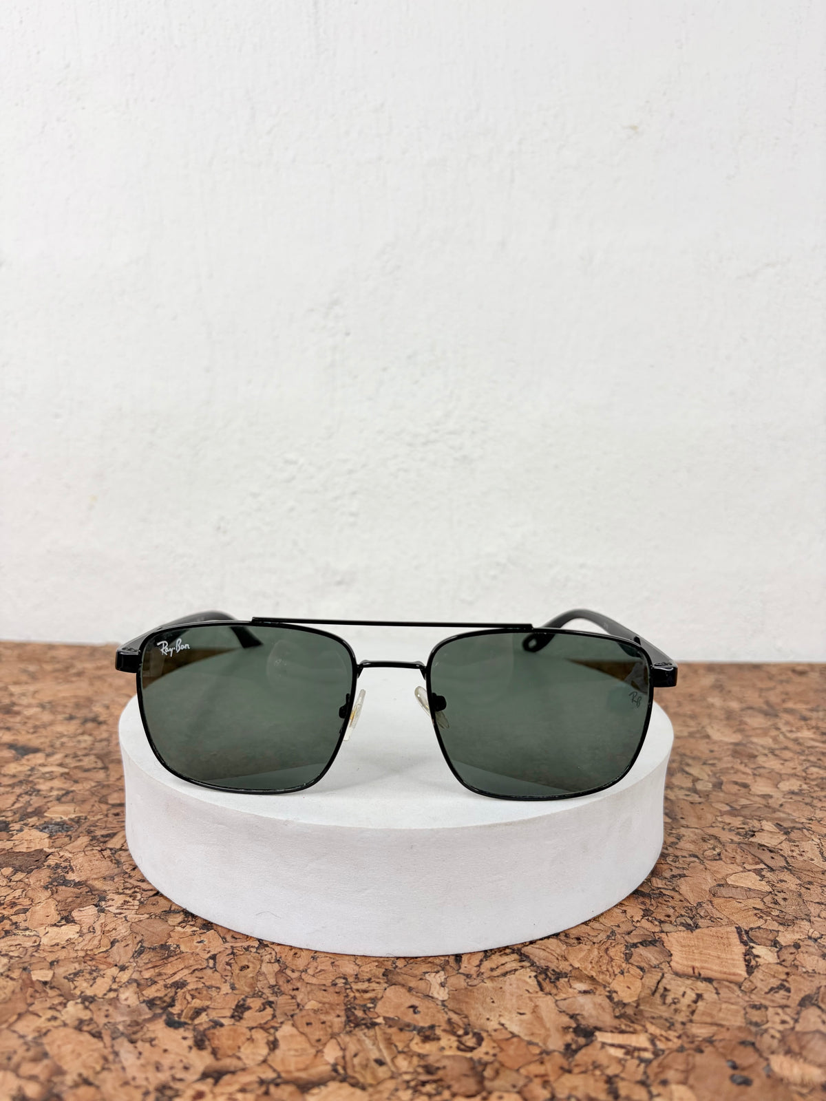Ray-Ban aviator sunglasses with dark green lenses and black frames, displayed on a white cylindrical riser over a cork surface against a white wall.