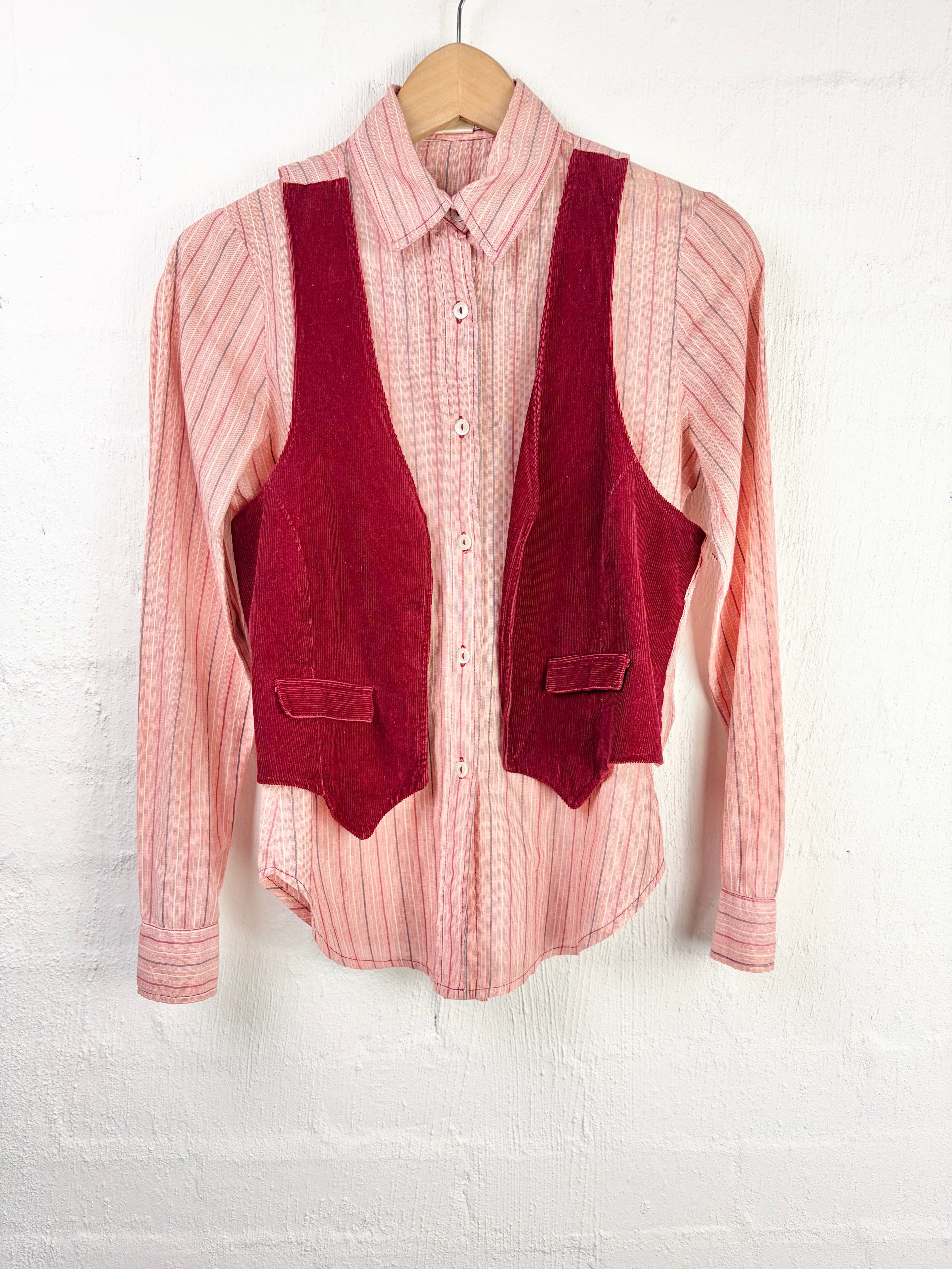 A pink and white vertically striped long-sleeve button-down shirt with a dark red corduroy vest layered over it, hanging on a wooden hanger against a white painted brick wall.