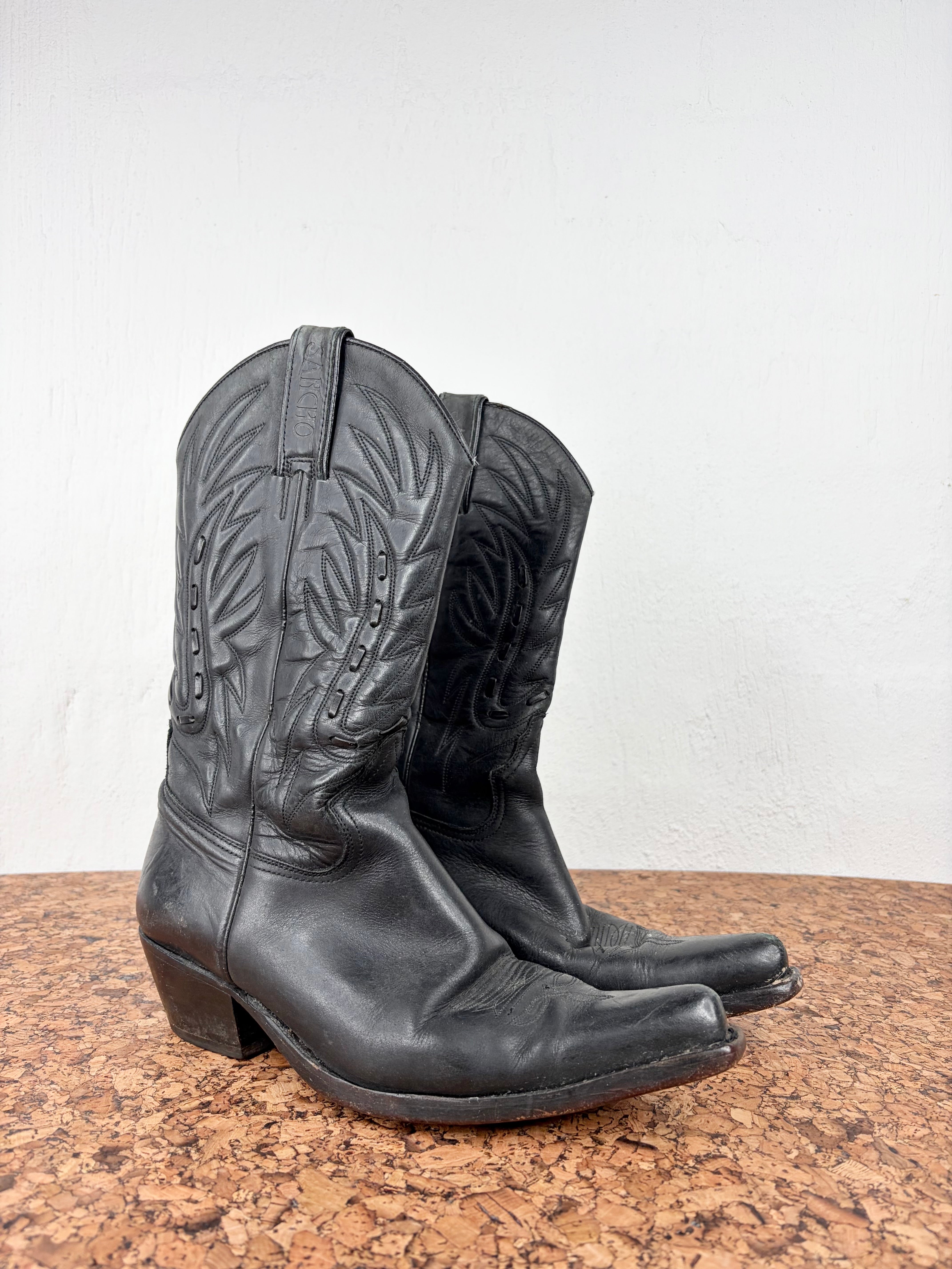 A pair of black leather cowboy boots with decorative western stitching, shown on a cork surface against a plain white wall.
