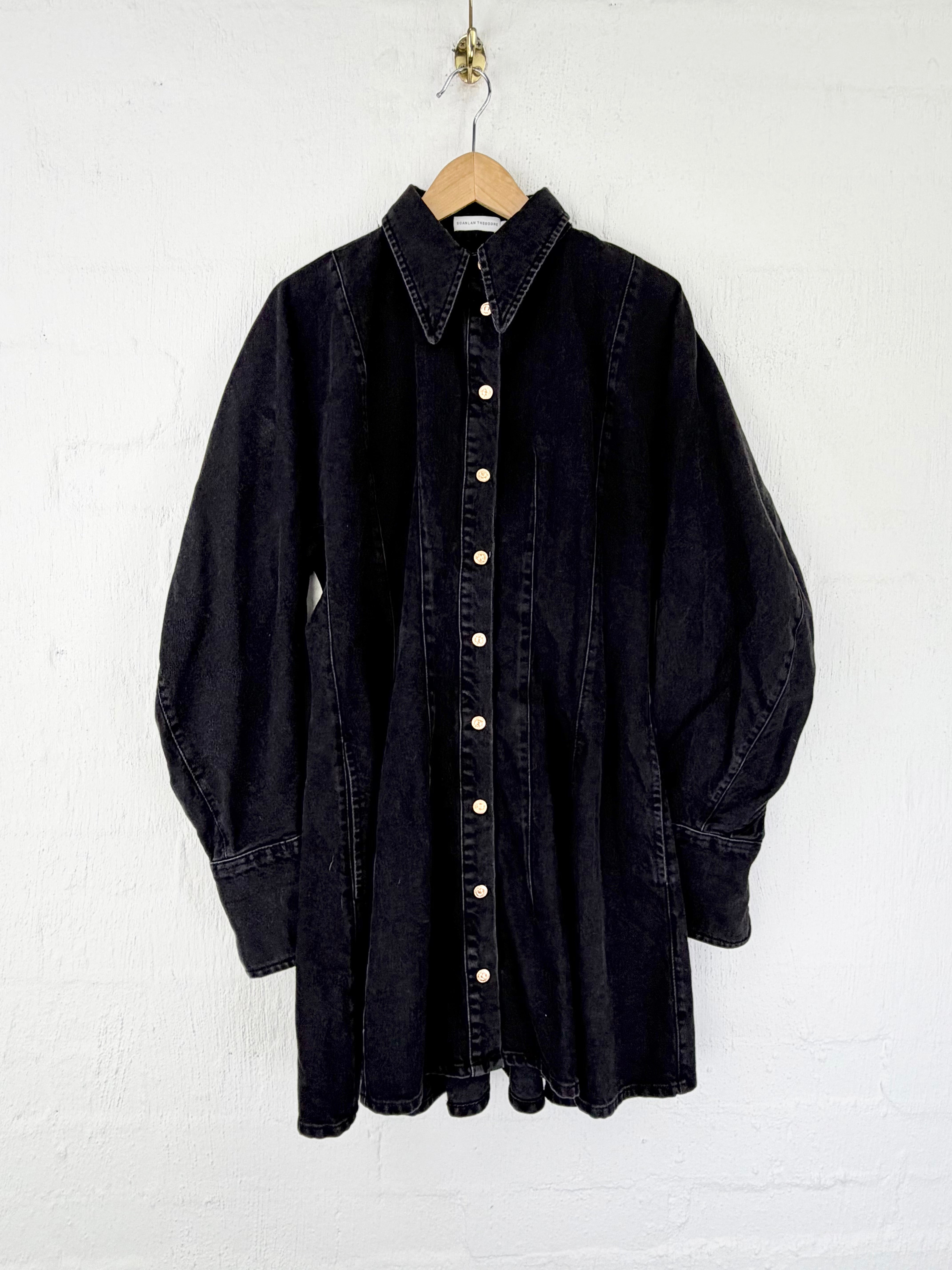 A black denim shirt dress with a pointed collar and front button closure, hanging on a wooden hanger against a white textured wall.