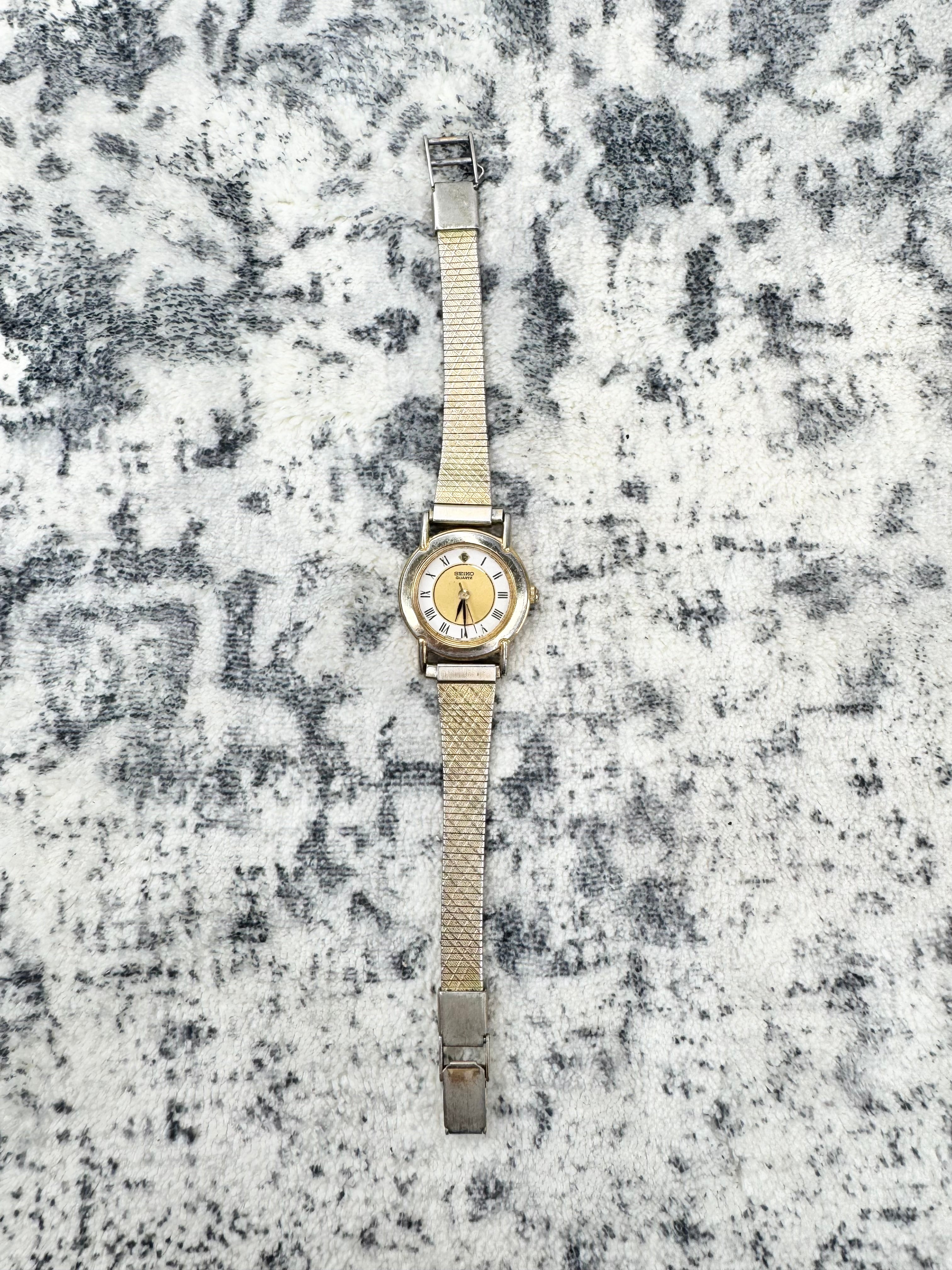 A vintage-style wristwatch with a gold-tone case and a beige mesh strap, displayed vertically on a textured gray and white patterned fabric background.