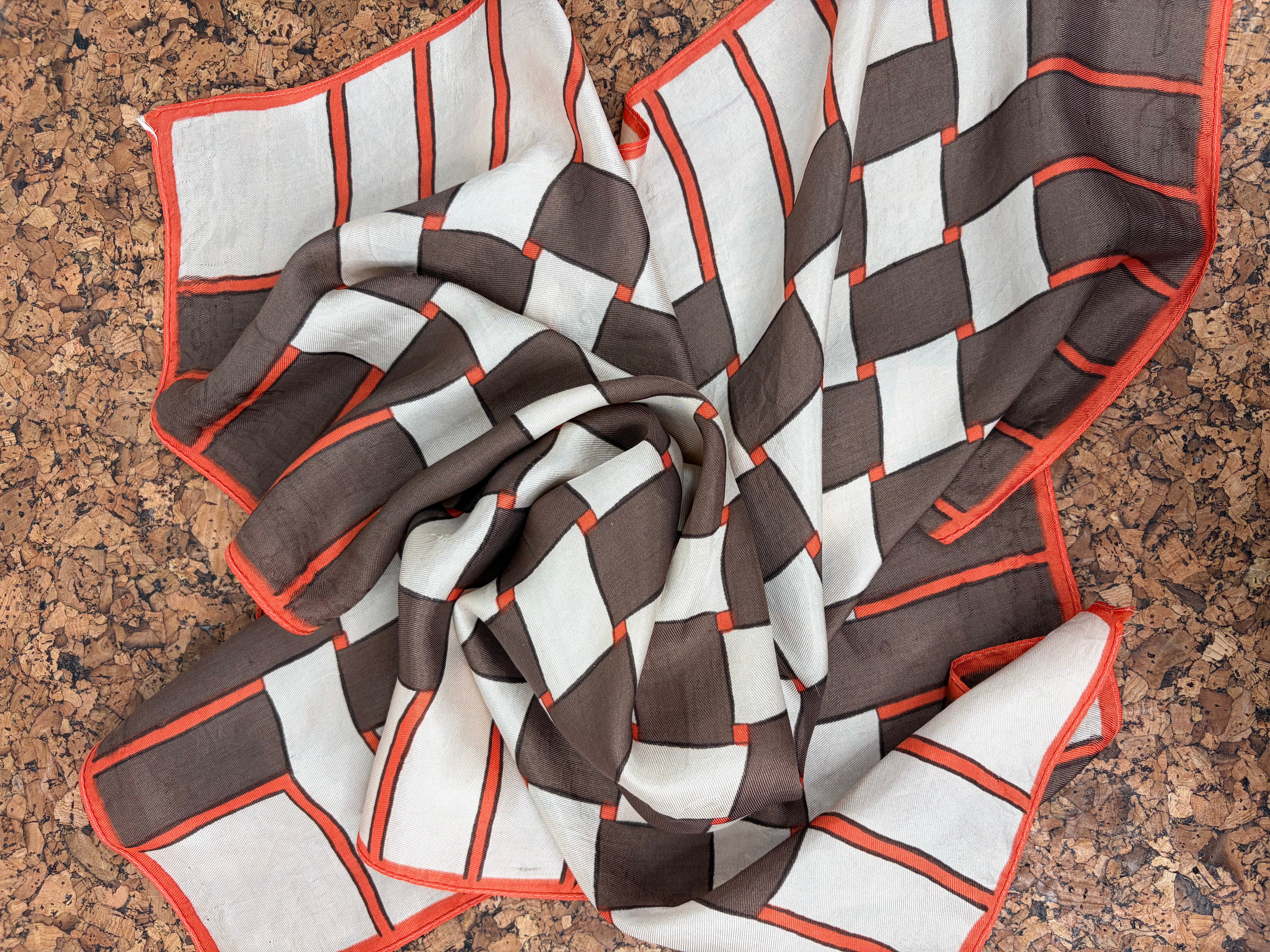 A square scarf with a geometric pattern of brown and white blocks outlined in orange-red, draped on a brown textured surface.