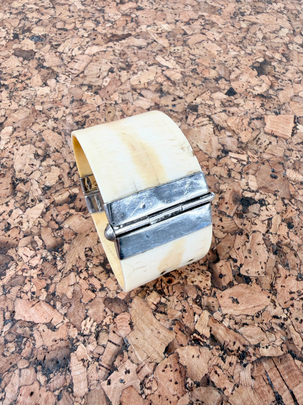 A wide cuff bracelet made of natural cream-colored bone or horn with a polished silver metal hinge clasp, resting on a textured cork surface.