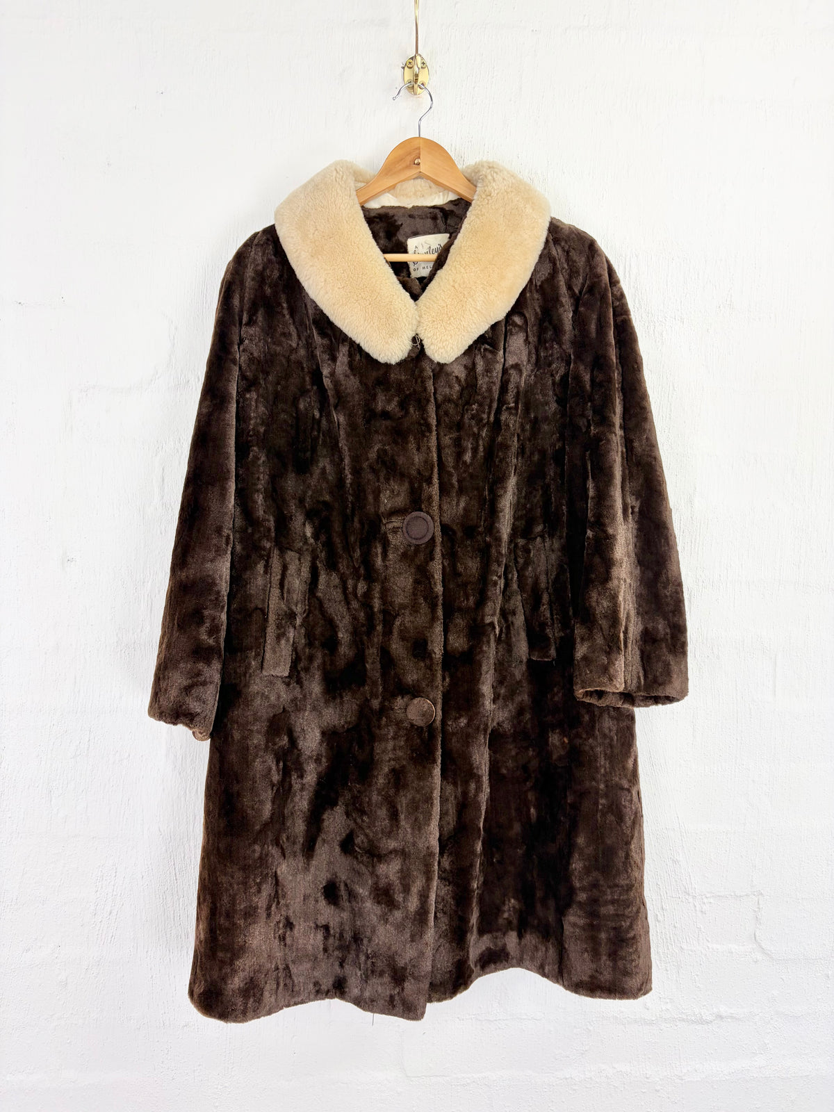 A dark brown plush faux fur coat with a cream-colored faux fur collar, hanging on a wooden hanger against a white painted brick wall.