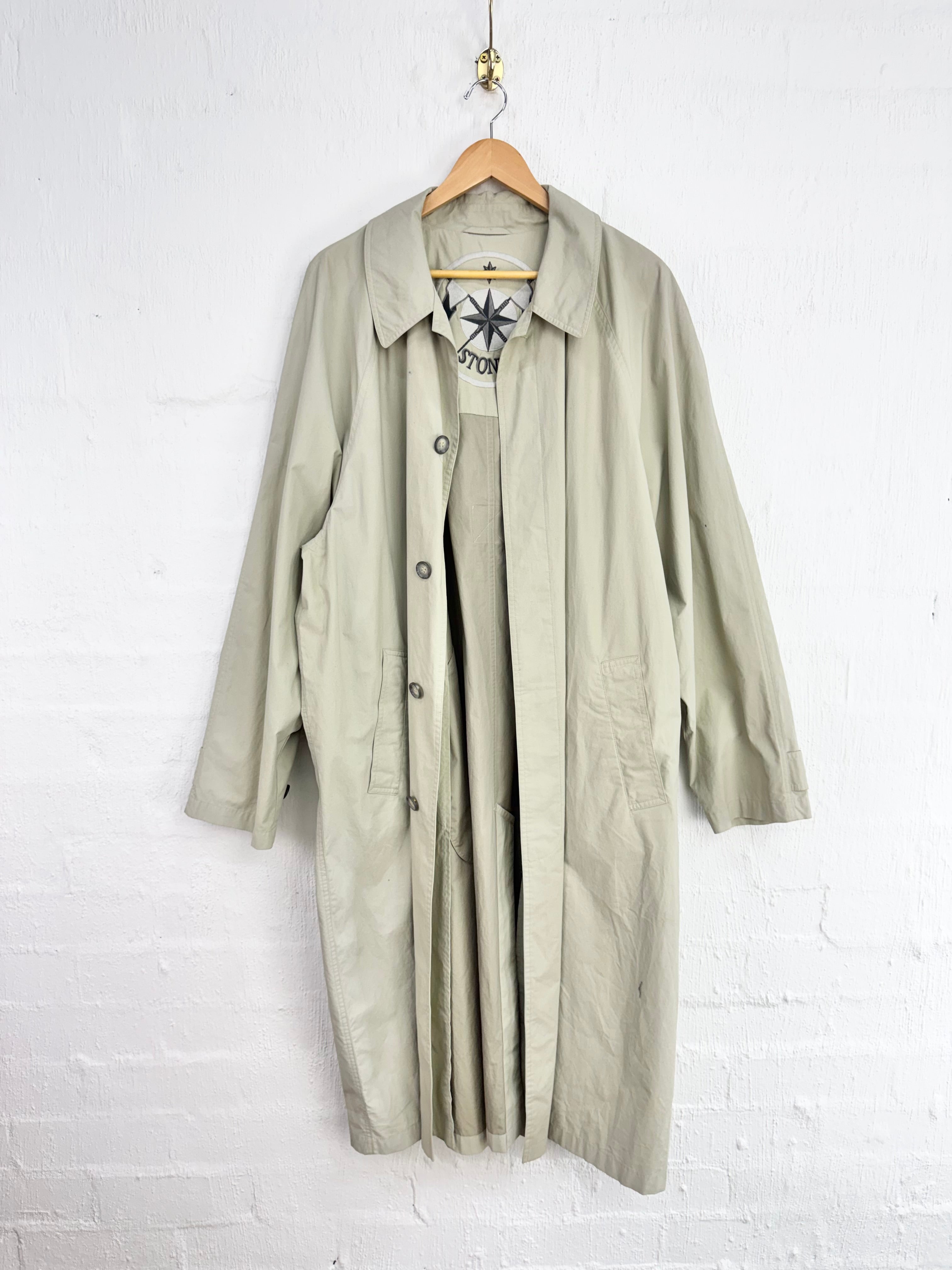 A light beige trench coat hanging on a wooden hanger against a white brick wall, layered over a matching beige dress with a black graphic print.