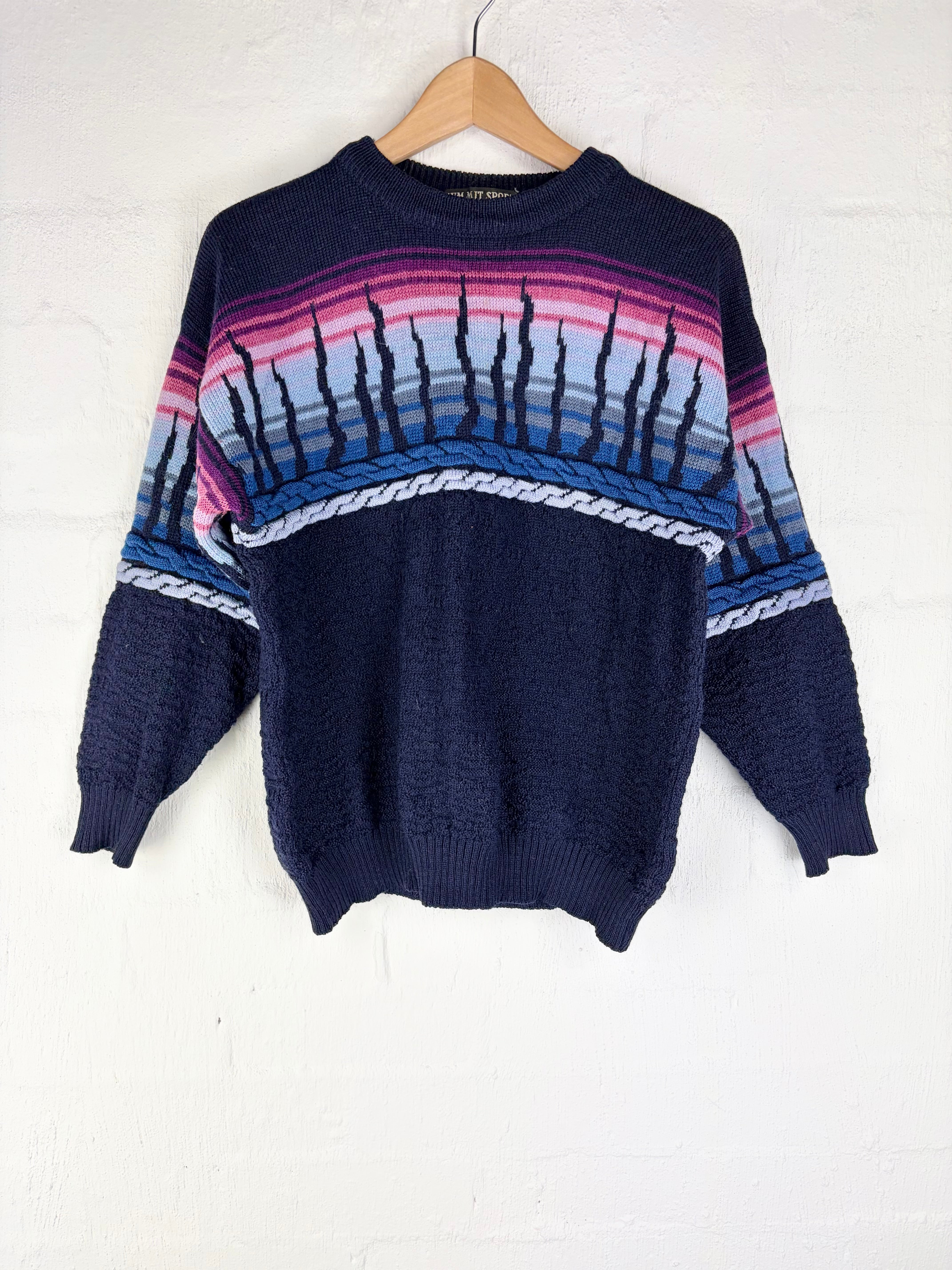 A vintage knit crewneck sweater in navy blue, featuring a colorful geometric pattern across the chest and sleeves with pink, purple, and light blue stripes, hanging on a wooden hanger against a white brick wall.