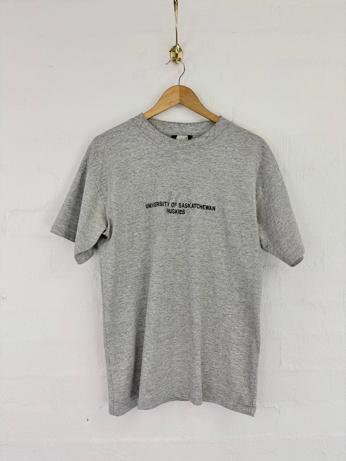 A heather gray short-sleeve t-shirt hanging on a wooden hanger against a white painted brick wall. The shirt features black printed text on the chest reading 'UNIVERSITY OF SASKATCHEWAN HANDBAL'.