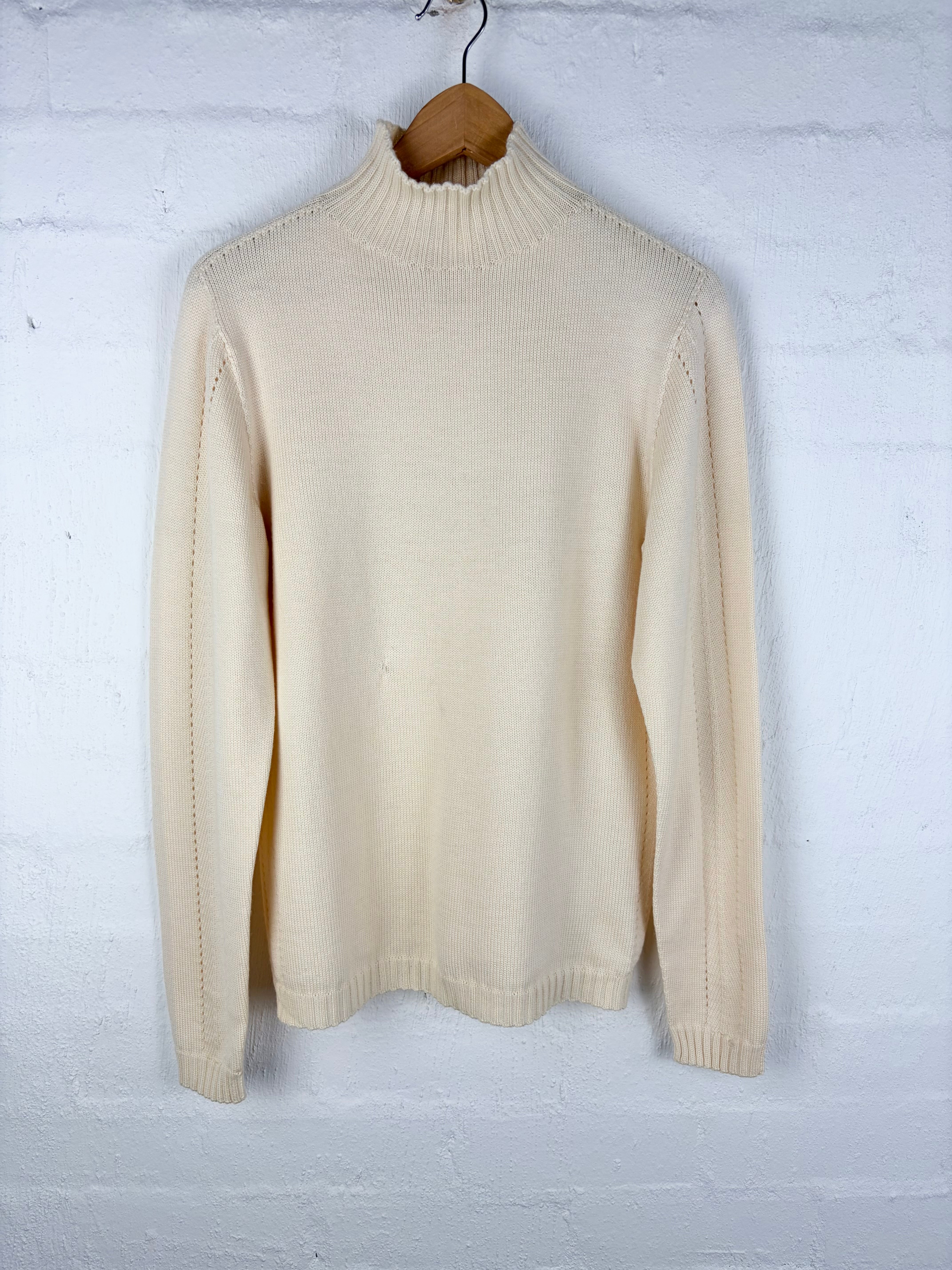 A cream-colored knit mock neck sweater hanging on a wooden hanger against a white painted brick wall.