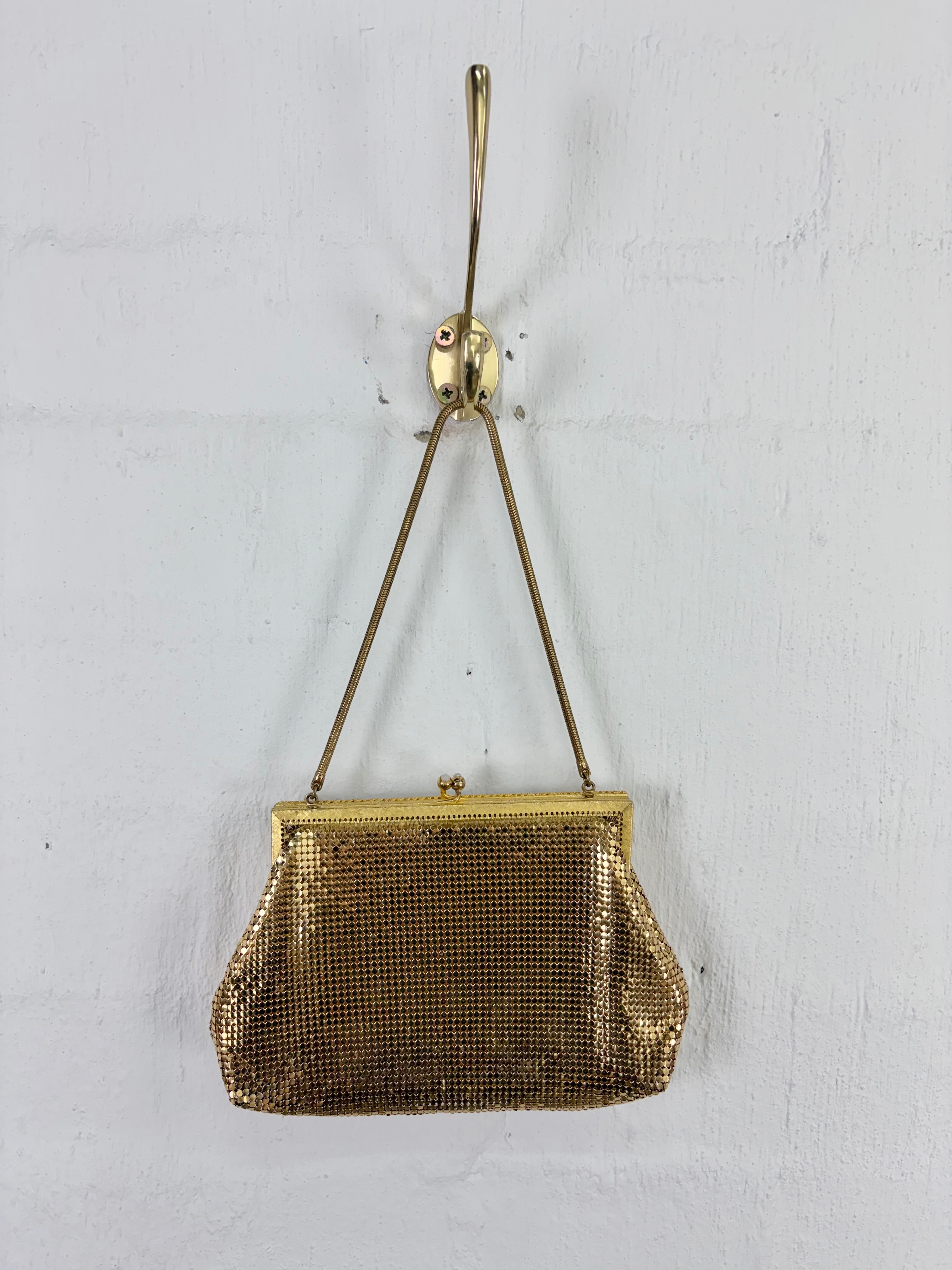 A vintage gold-tone mesh clutch purse with a gold-tone metal frame and shoulder strap, hanging from a brass wall hook against a white textured wall.