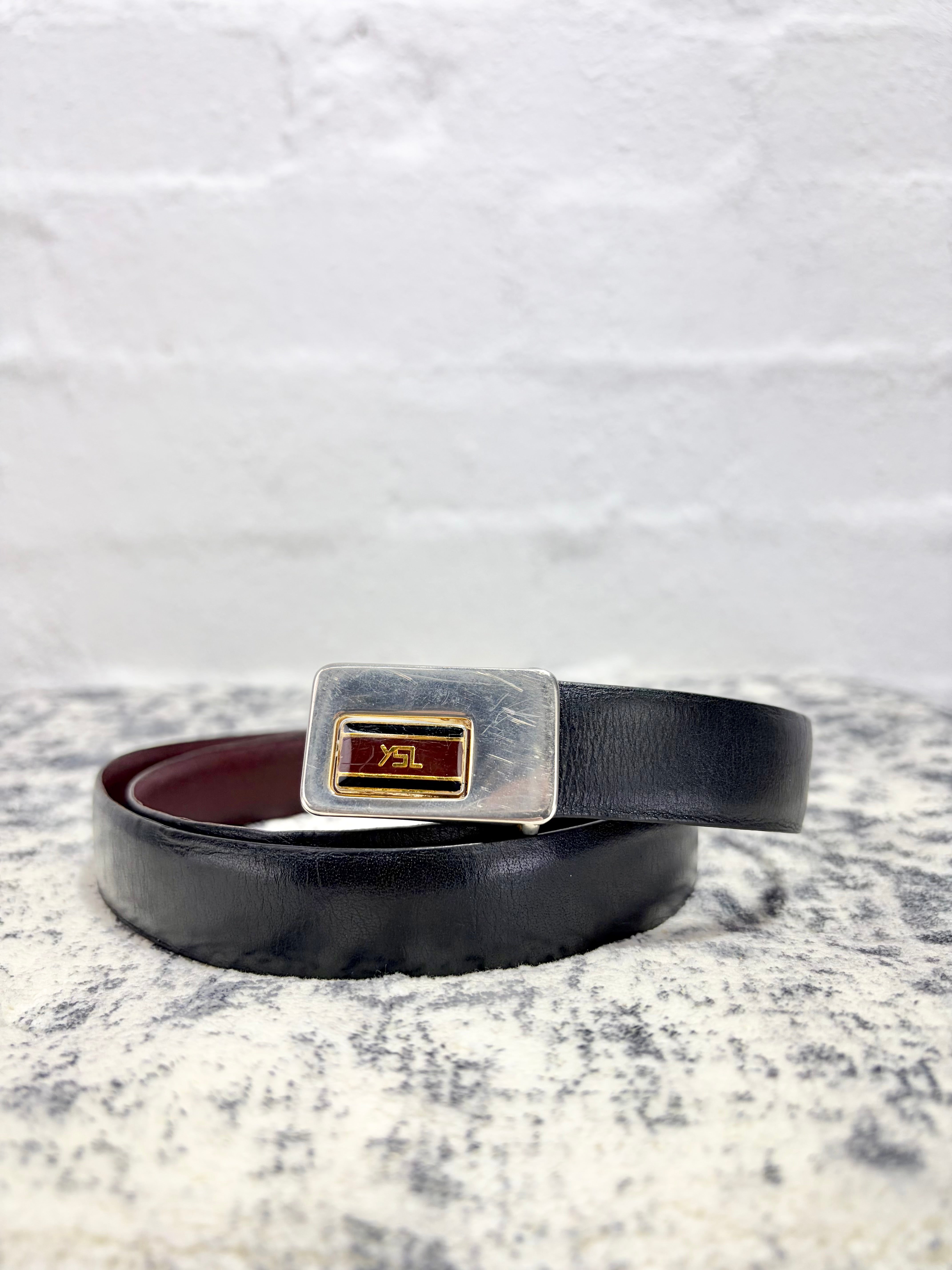 A black leather belt with a silver-toned rectangular buckle featuring a red and gold center plate, coiled on a textured gray surface against a white brick wall background.