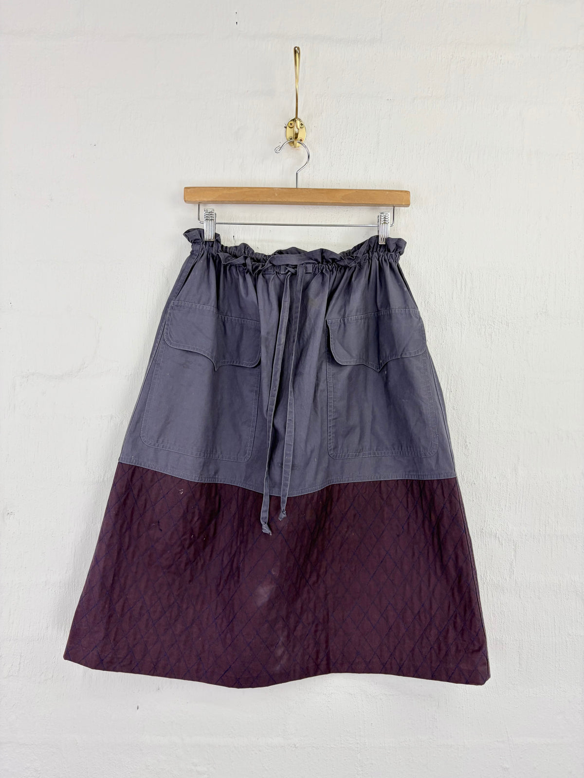 A two-tone A-line skirt hanging on a wooden hanger against a white painted brick wall. The top half is a muted slate gray, and the bottom half is a deep plum purple.