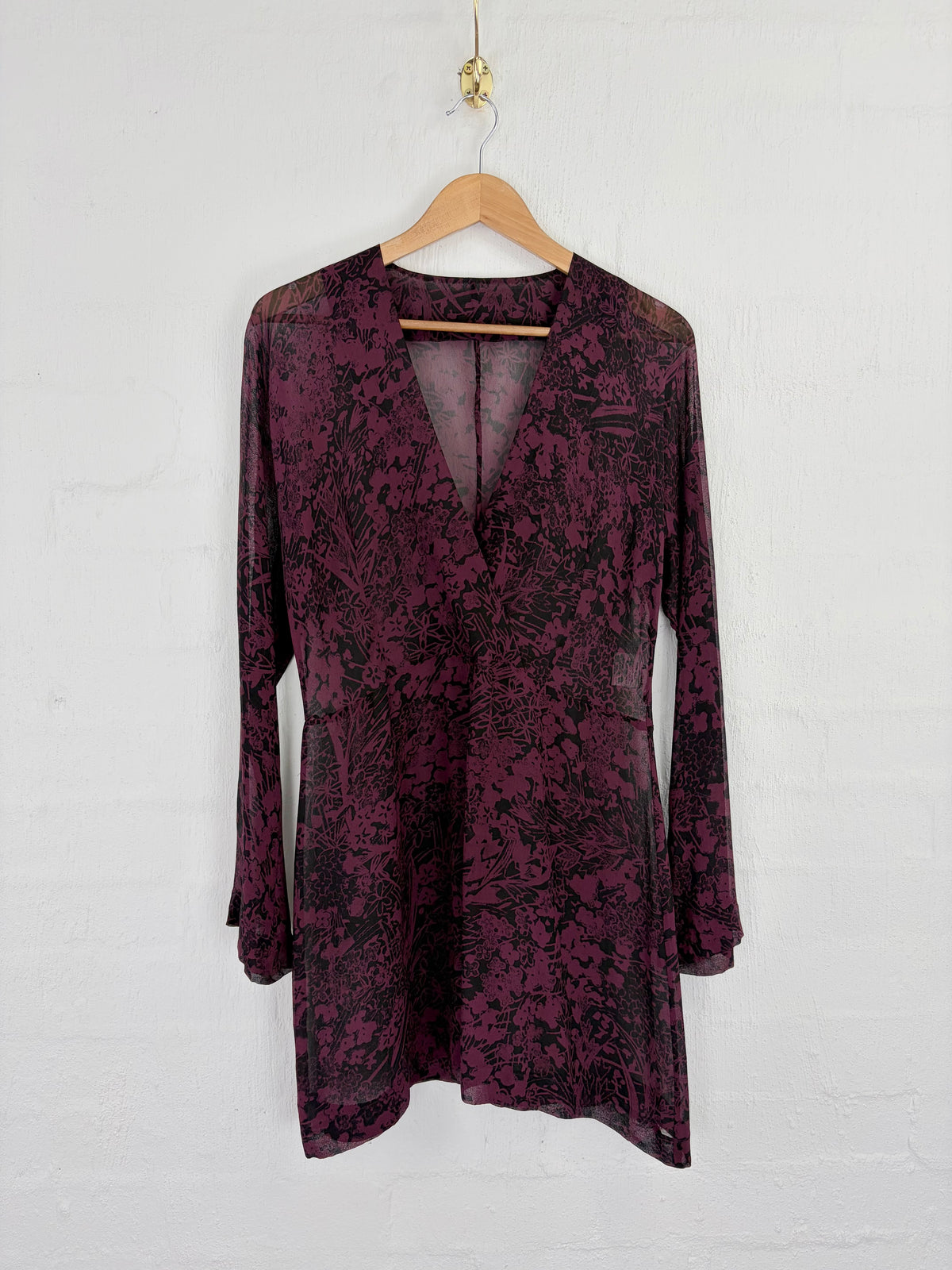 A long-sleeve tunic dress in a dark purple and black floral jacquard pattern, hanging on a wooden hanger against a white painted brick wall.