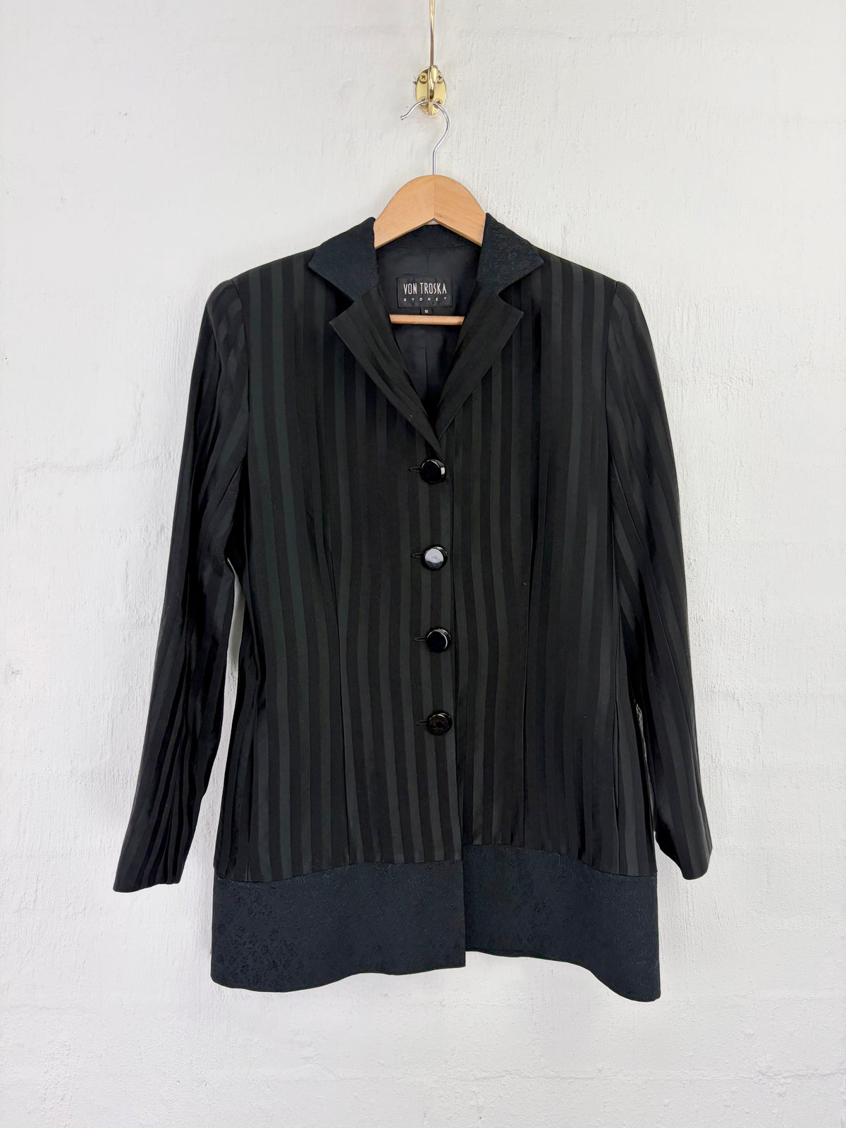A black long-sleeve blazer with vertical stripes and a button-front closure, hanging on a wooden hanger against a white wall. The brand label reads 'SUN TREES'.