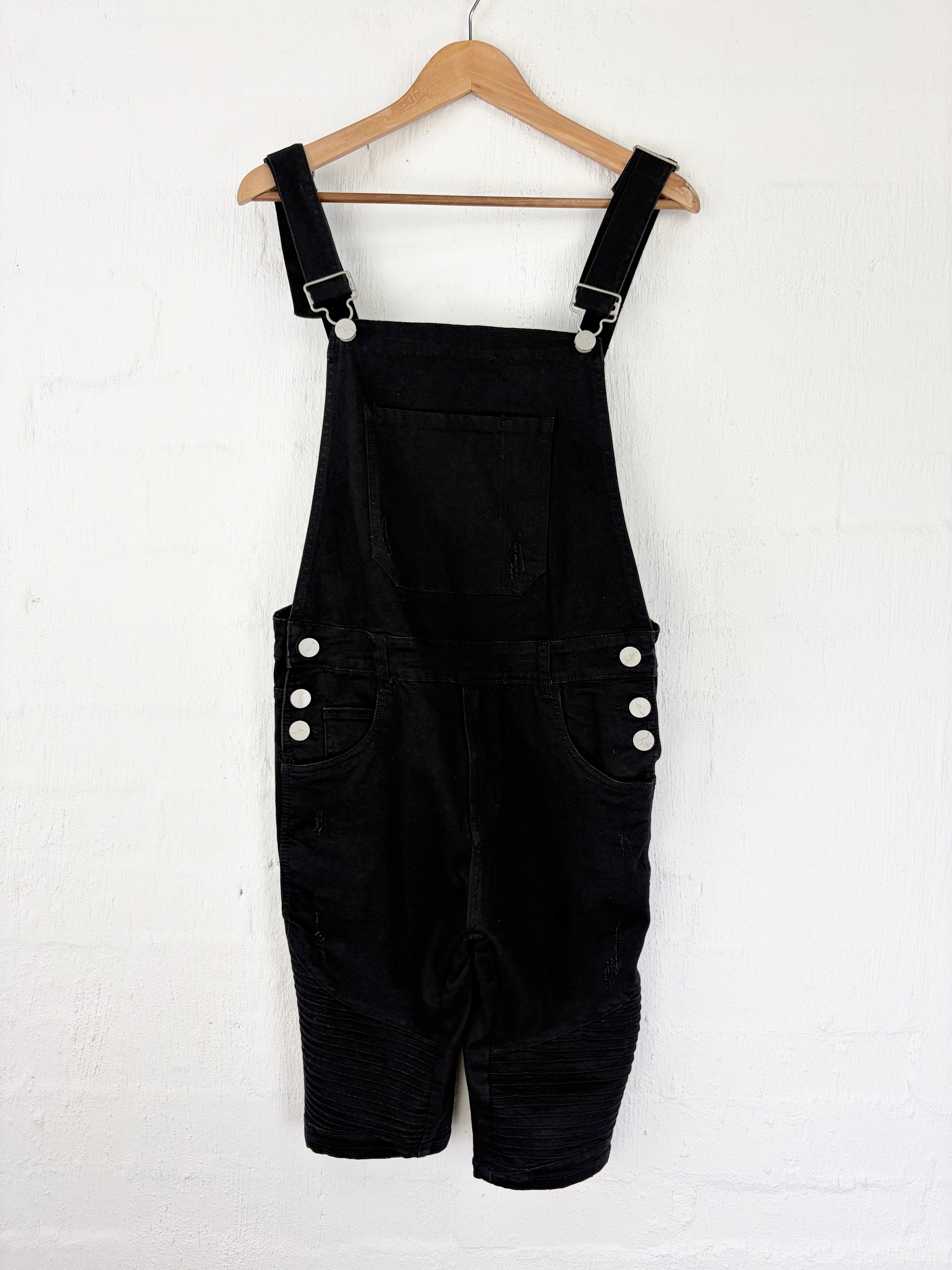 Black corduroy short overalls hanging on a wooden hanger against a white painted brick wall. The bib has a front pocket and adjustable shoulder straps with silver metal buckles, and the shorts feature side pockets with white buttons.