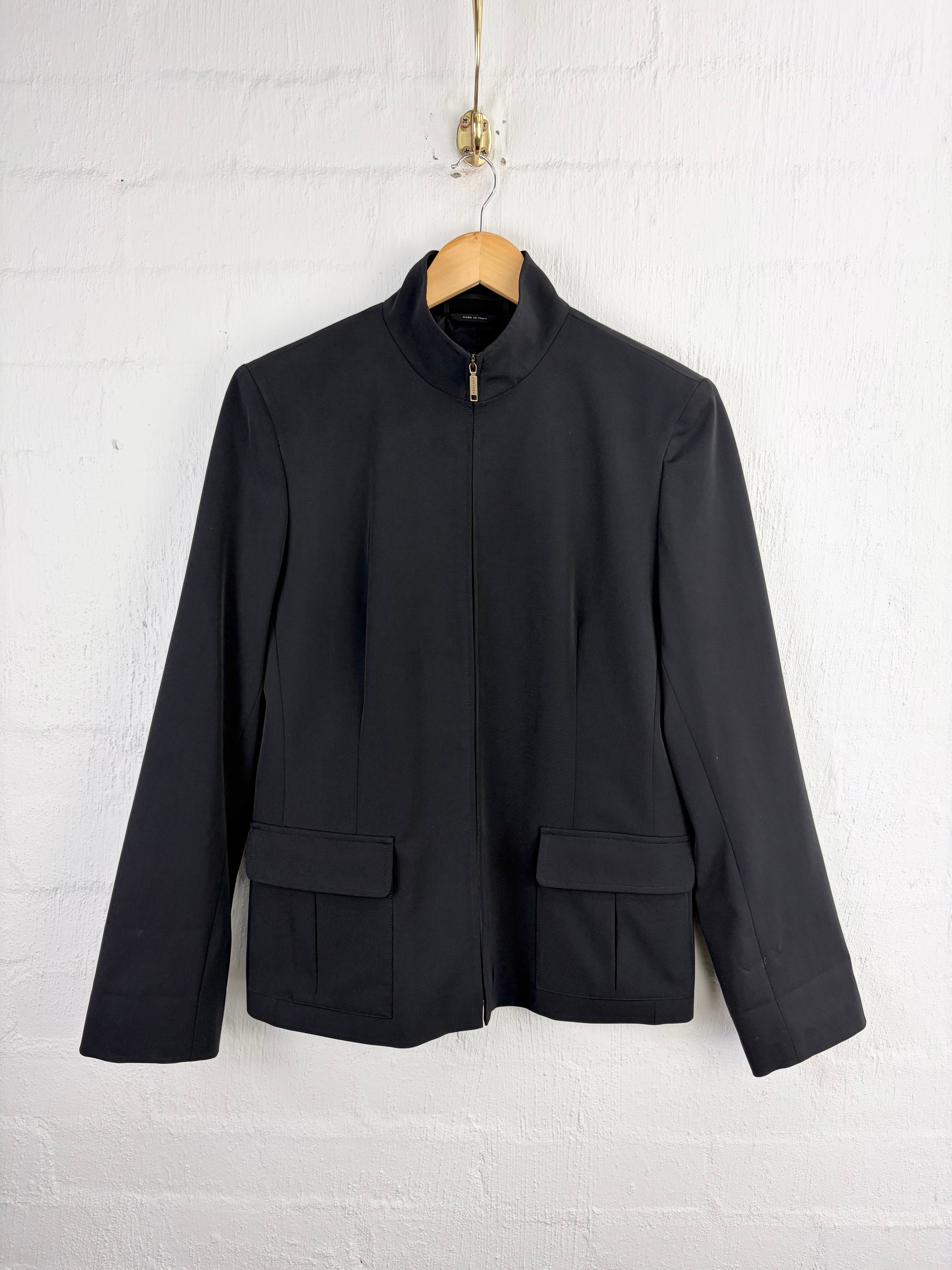 A black long-sleeve jacket with a stand collar and front zipper, hanging on a wooden hanger against a white painted brick wall.