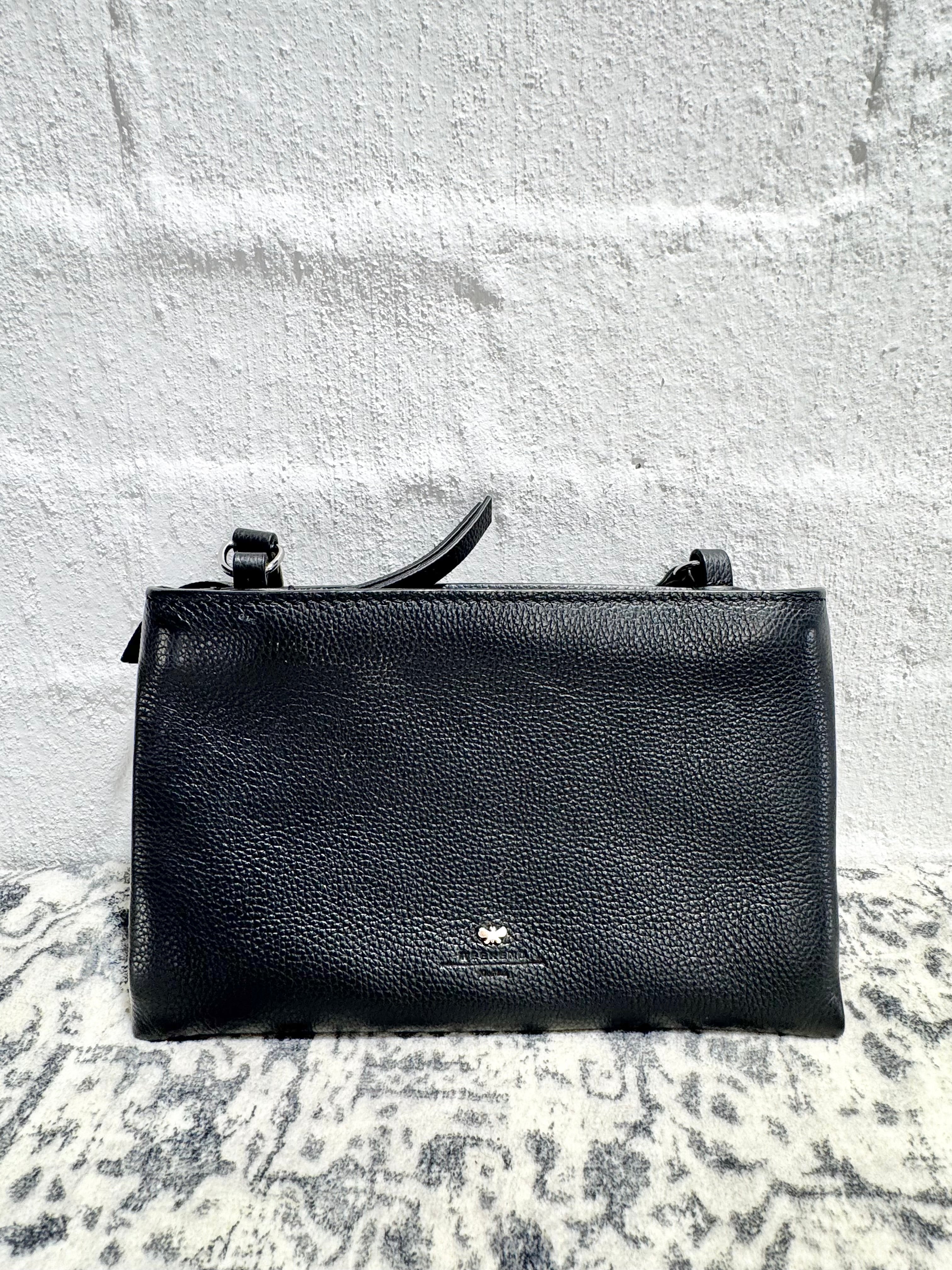 A black pebbled leather clutch bag with a top handle, resting on a gray and white patterned rug against a white brick wall.