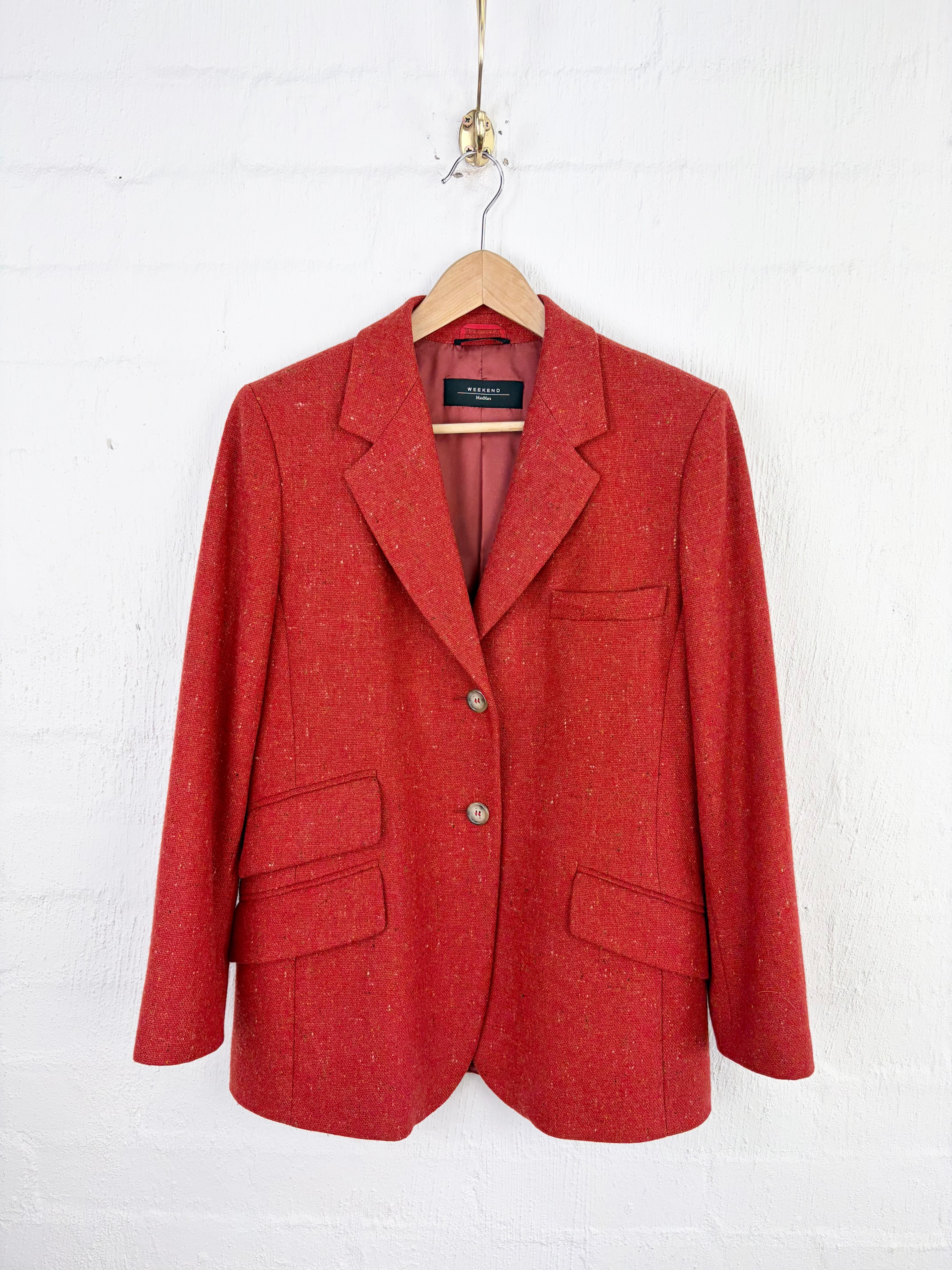 A red textured wool blazer jacket hanging on a wooden hanger against a white painted brick wall.