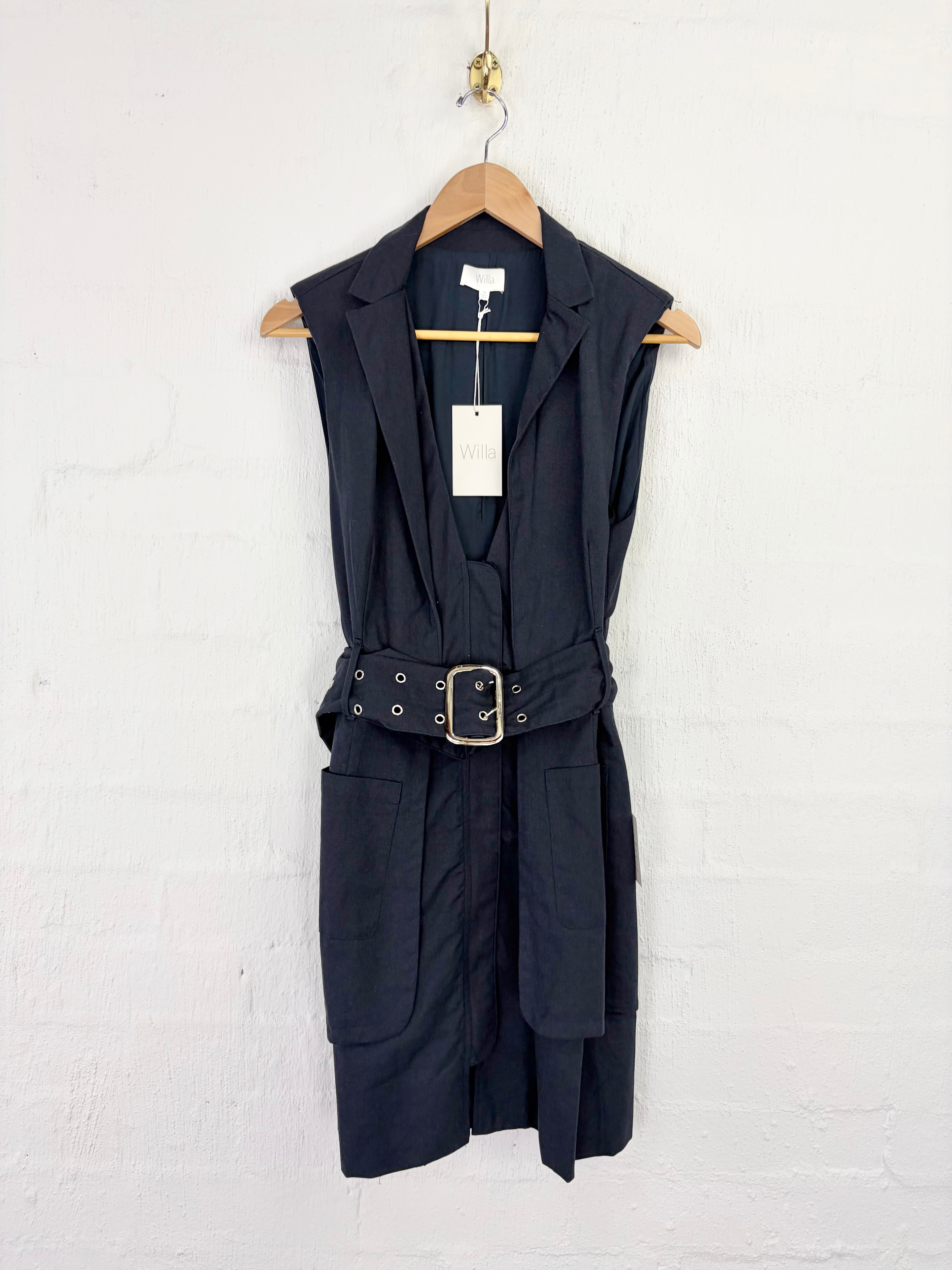 A sleeveless black belted vest dress with notched lapels and front patch pockets, hanging on a wooden hanger against a white painted brick wall.