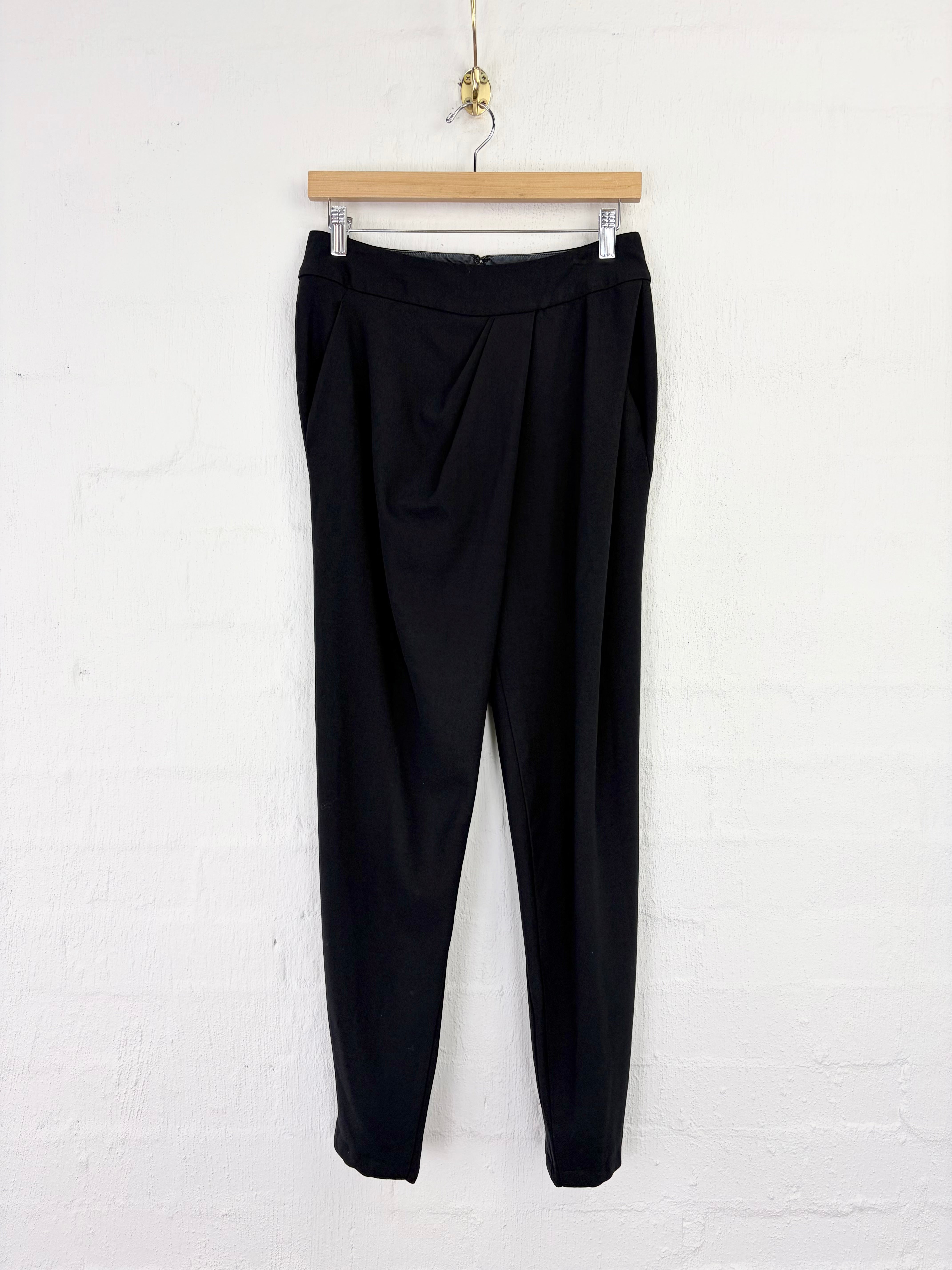 Black tailored trousers hanging from a wooden hanger against a white painted brick wall.