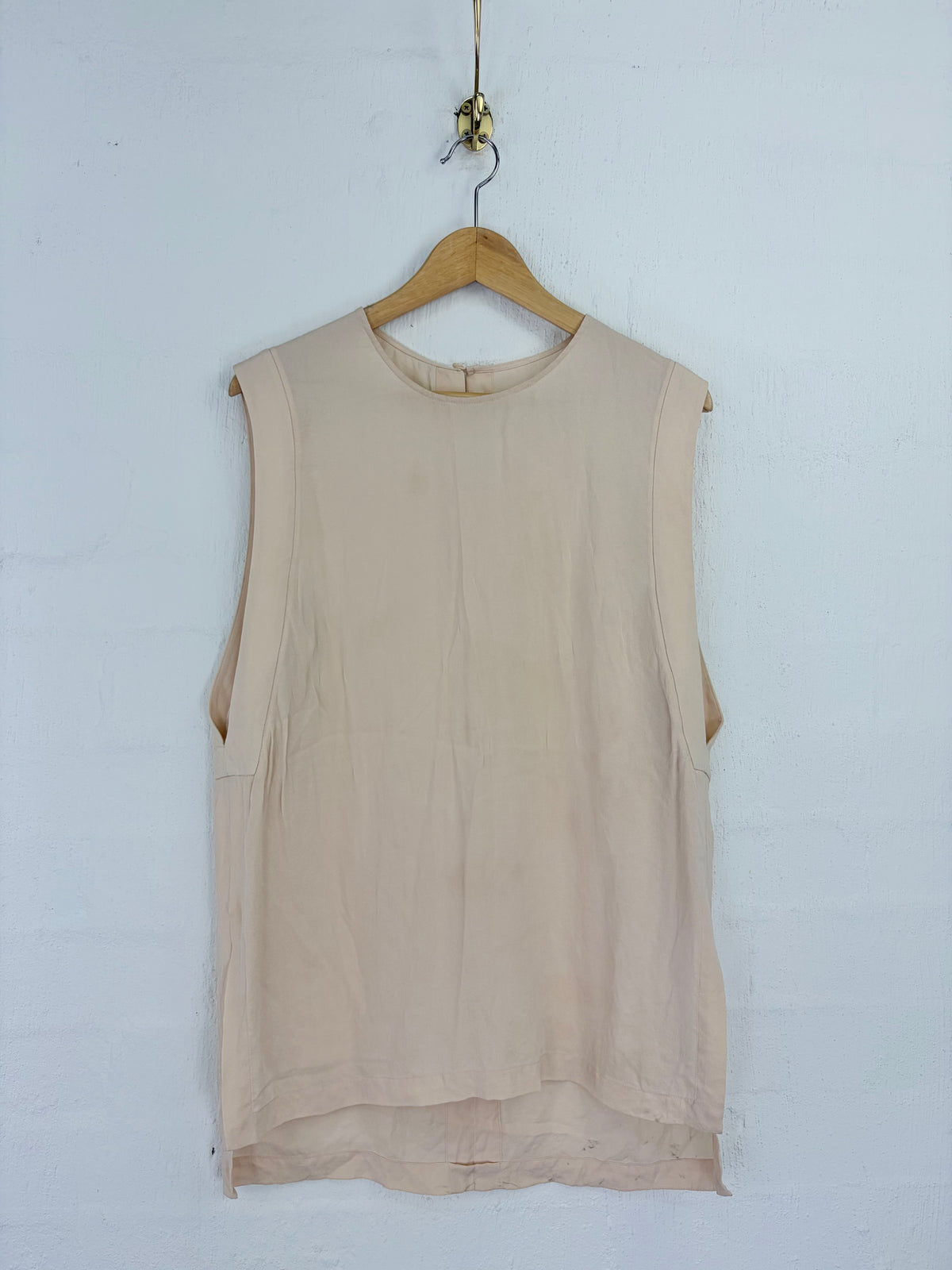 A sleeveless beige top hanging on a wooden hanger against a white painted brick wall.