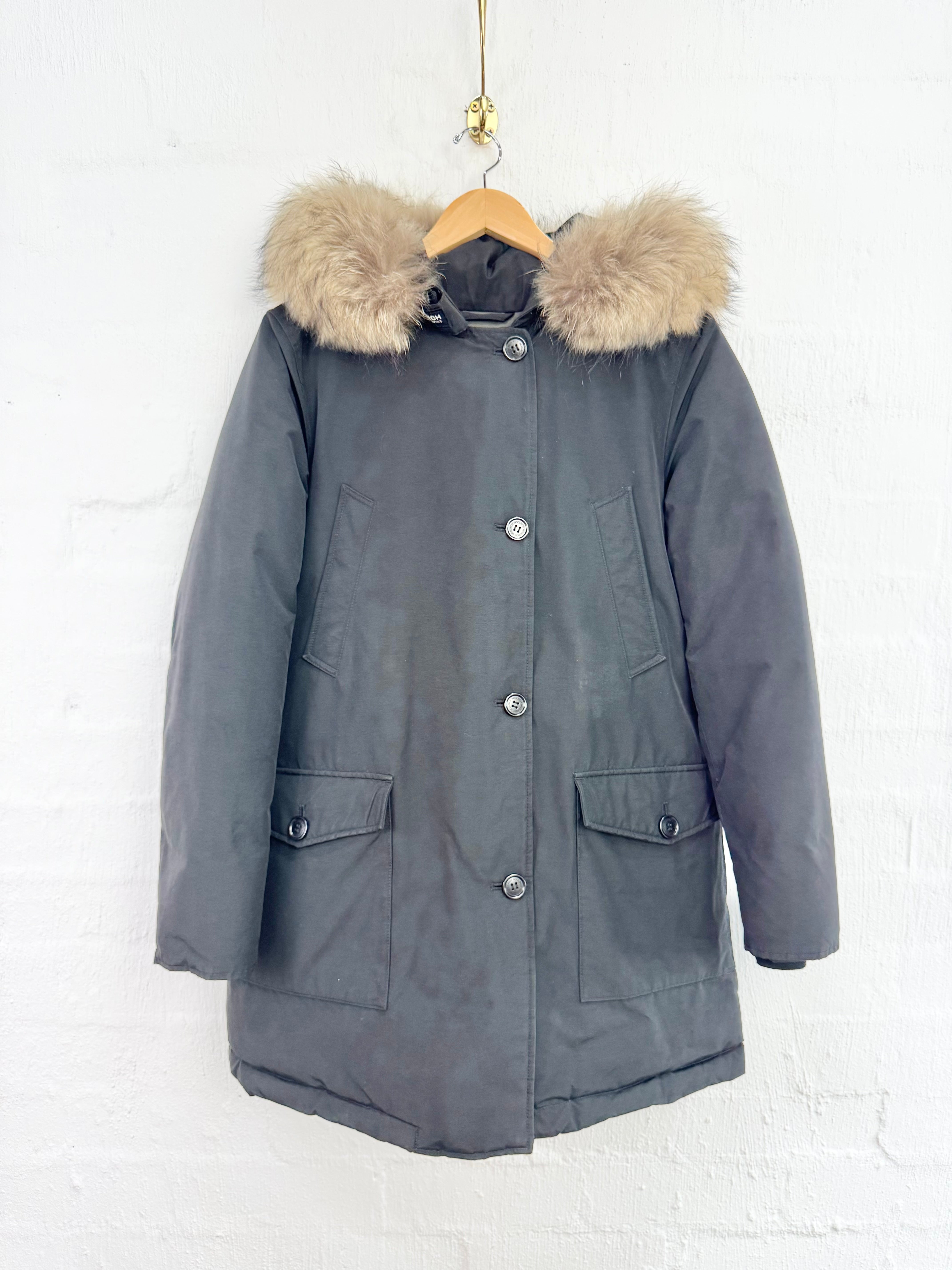 A dark gray hooded puffer parka jacket with a beige faux-fur trimmed hood, hanging on a wooden hanger against a white brick wall.