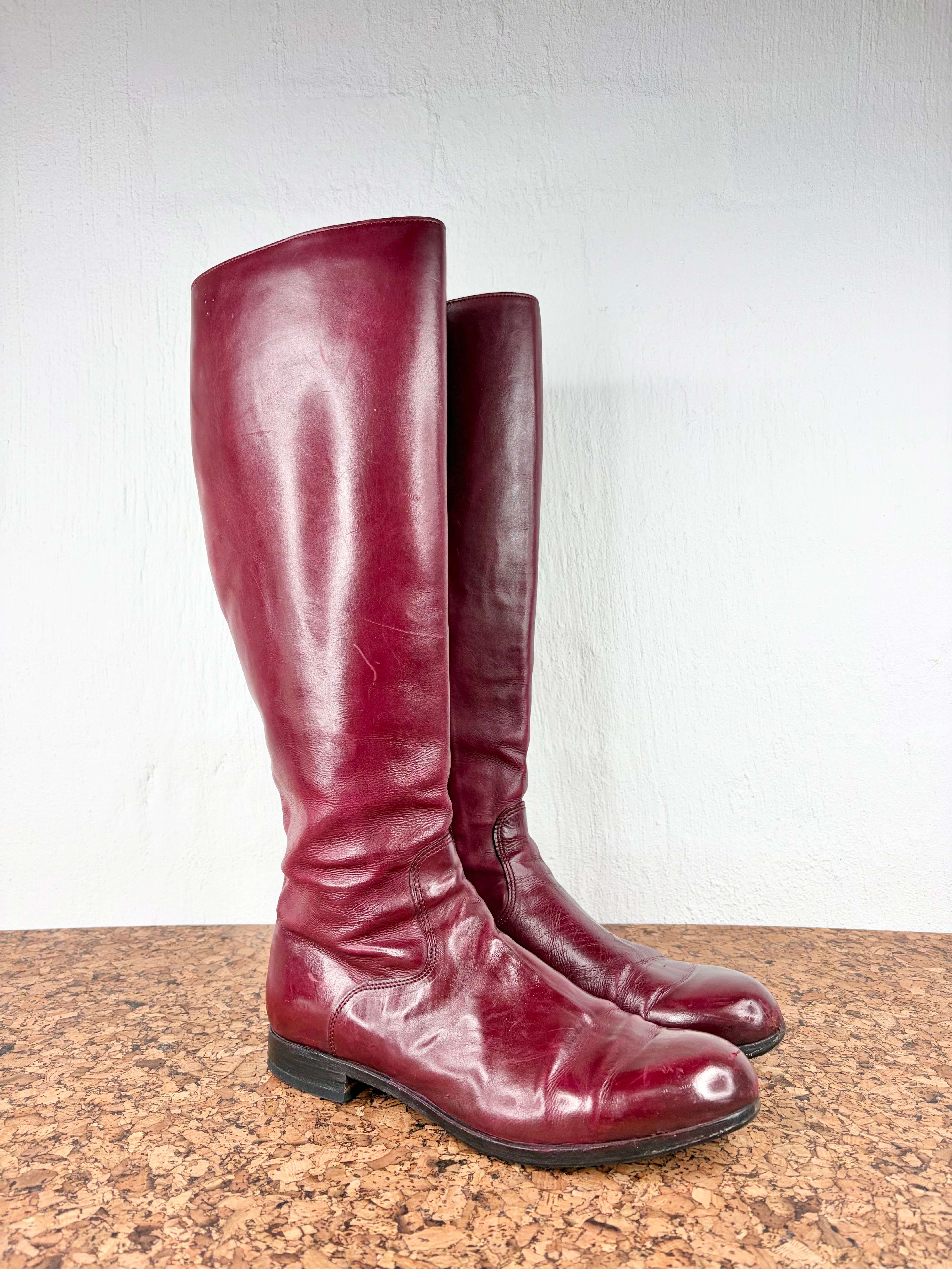 A pair of knee-high leather boots in a deep burgundy red, standing on a brown cork surface against a white wall.