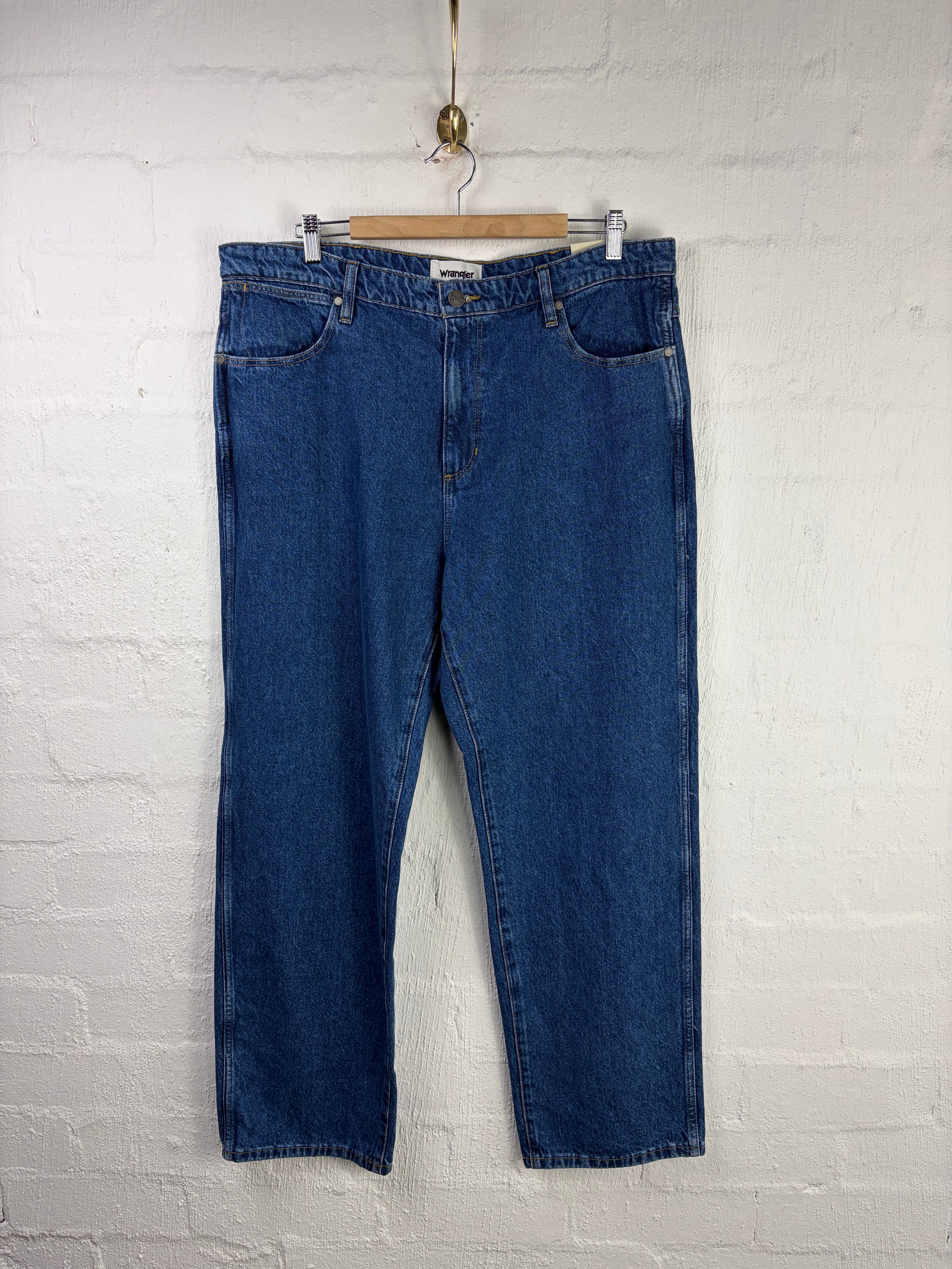 A pair of medium-wash blue denim jeans hanging from a wooden hanger against a white painted brick wall.