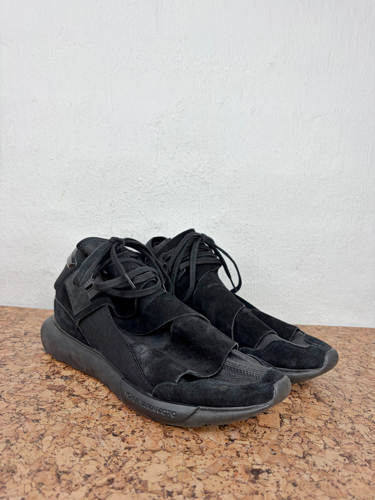 A pair of black high-top sneakers with a textured suede and mesh upper, displayed on a cork surface against a white wall.